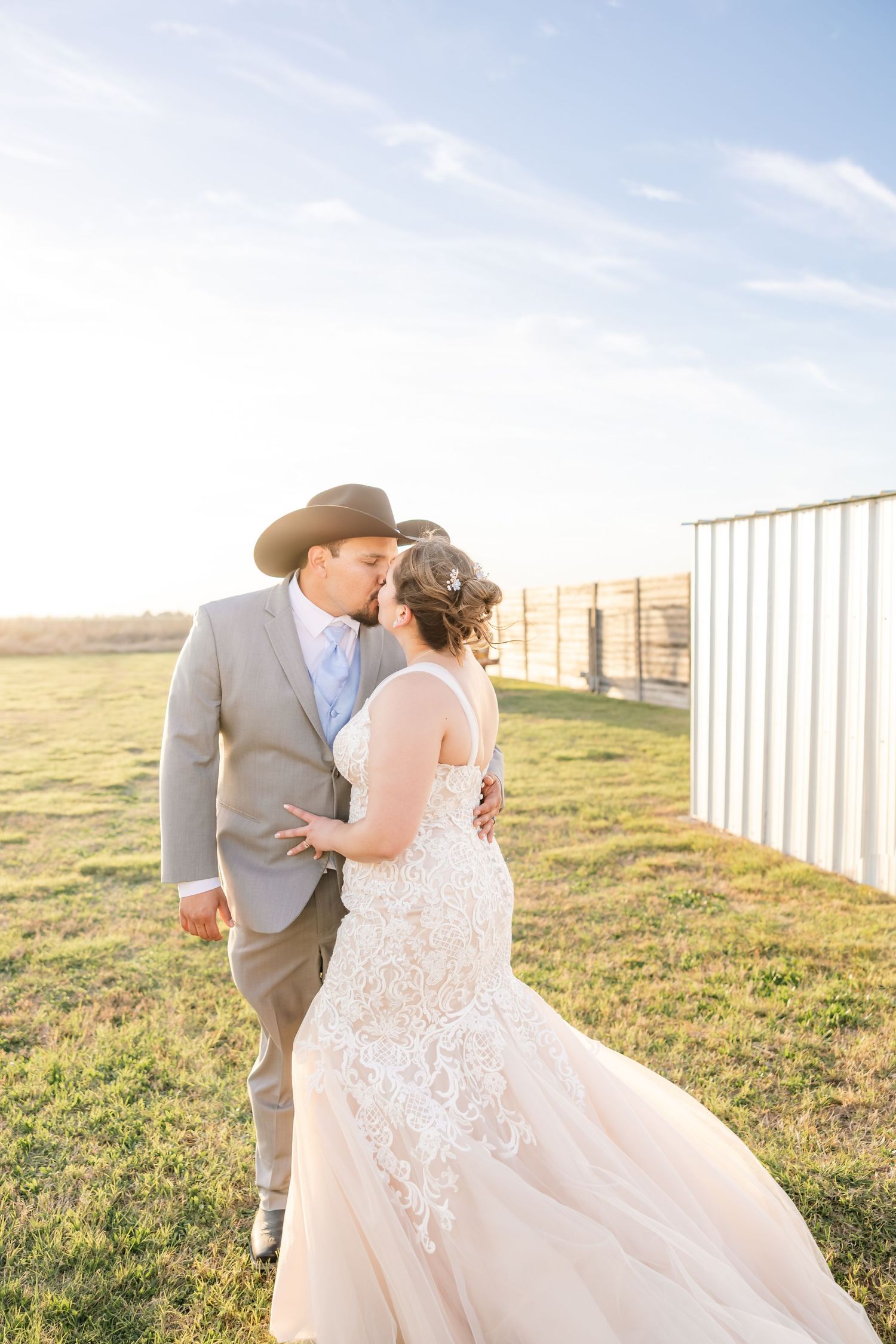 Bride and groom share a romantic kiss in a sunlit field, groom wearing a black cowboy hat and gray suit.