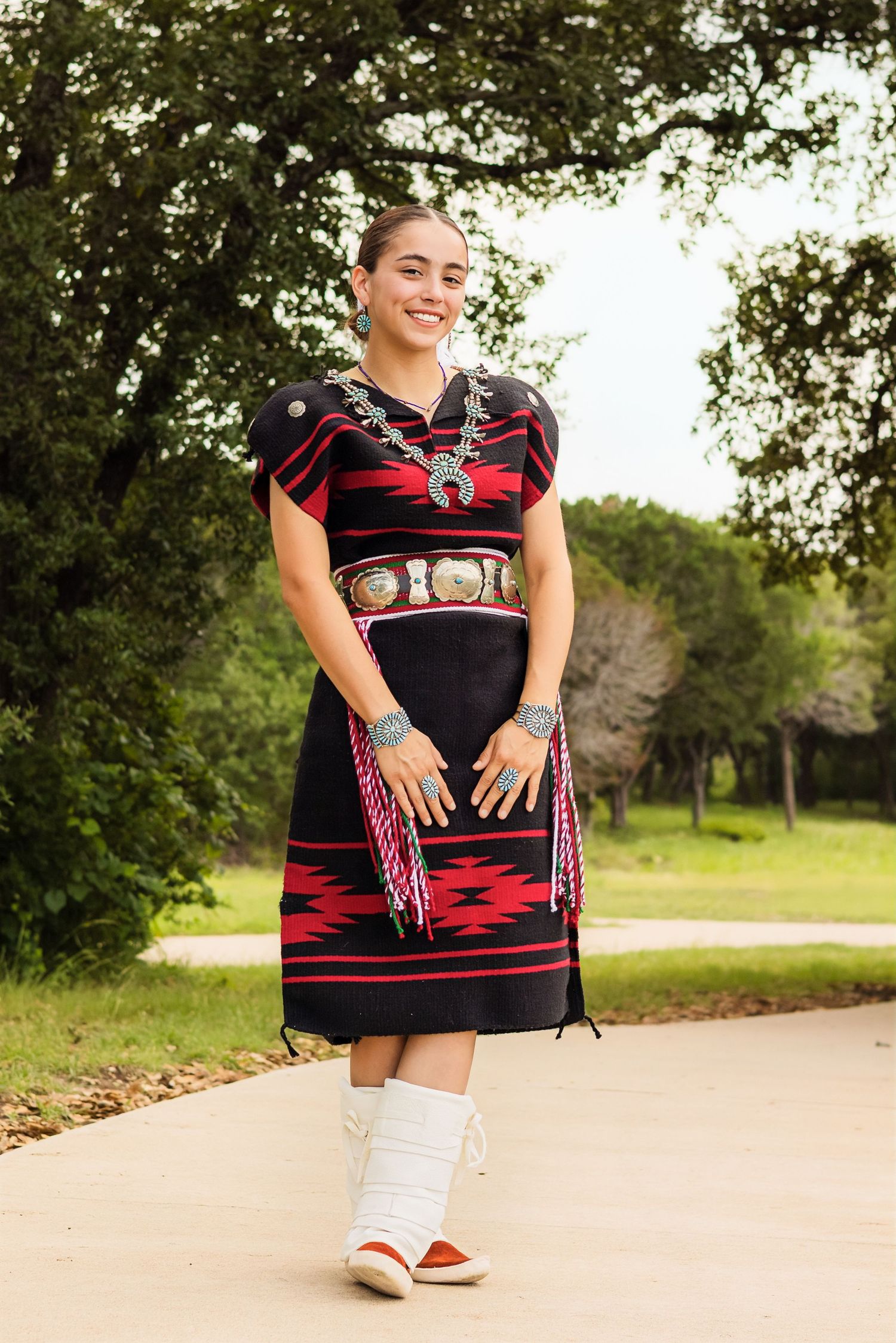 Young woman in traditional Native American dress with silver jewelry and white moccasin boots outdoors.