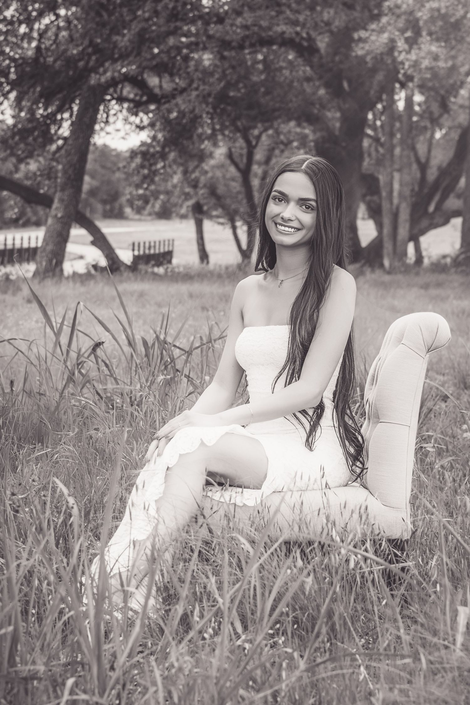 Young woman in white dress and boots sits smiling in a chair outdoors among tall grass and trees in black and white.