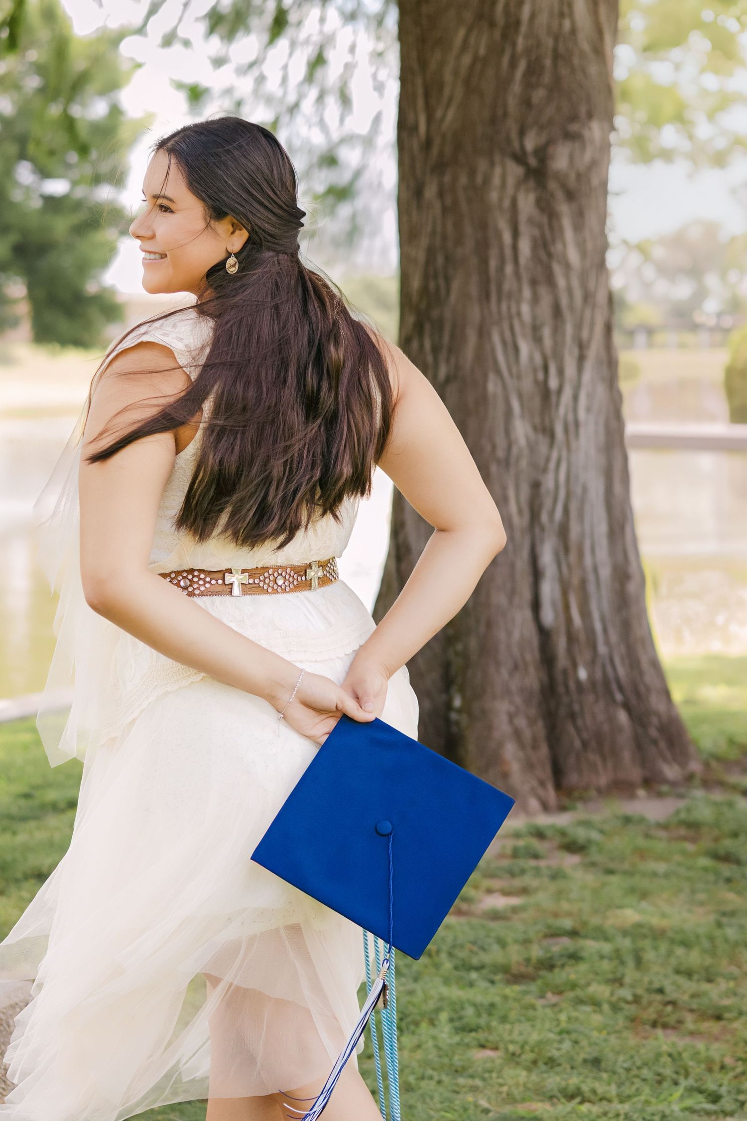 Smiling graduate in white dress holding blue mortarboard cap near a tree outdoors.