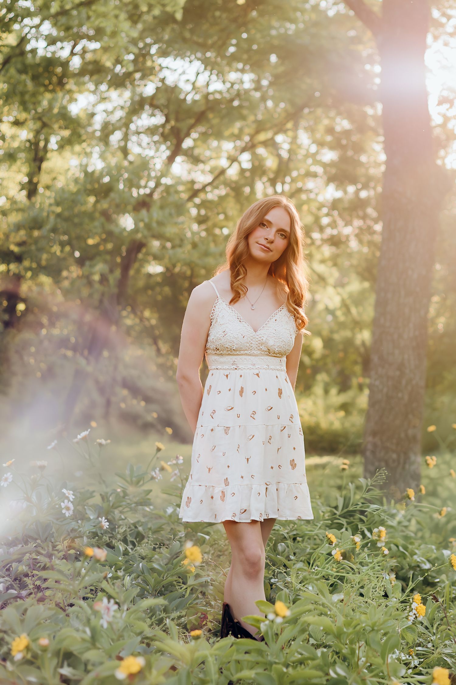 Young woman in white floral sundress standing among wildflowers in sunlit forest clearing during golden hour.