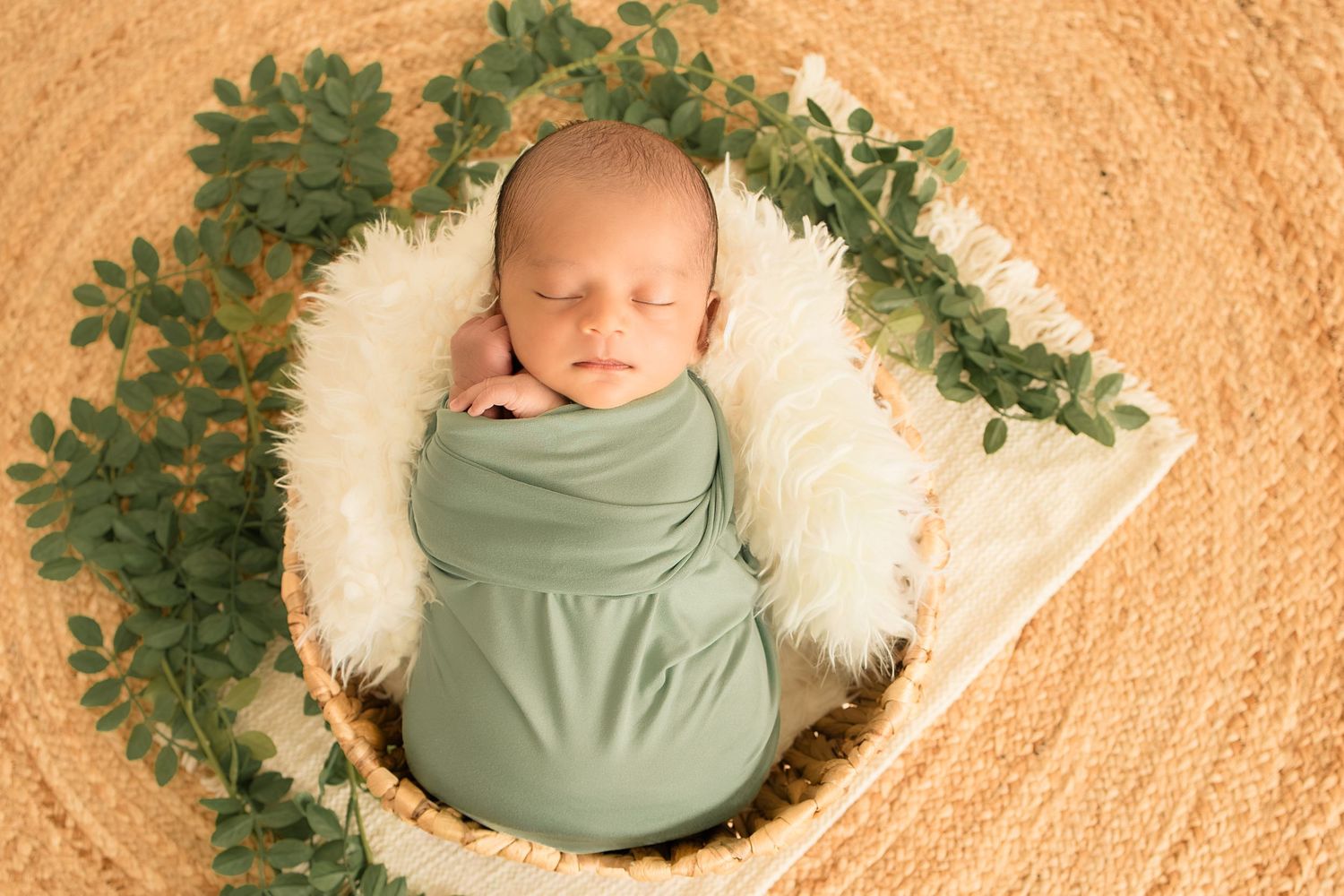 Newborn baby swaddled in sage green wrap sleeping peacefully in a wicker basket with white fur and green leaves.
