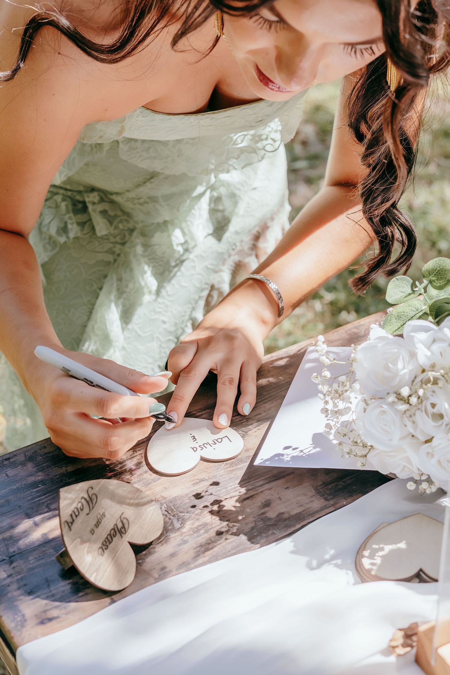 Bridesmaid in sage green dress signs wooden heart guest book at outdoor wedding reception table.