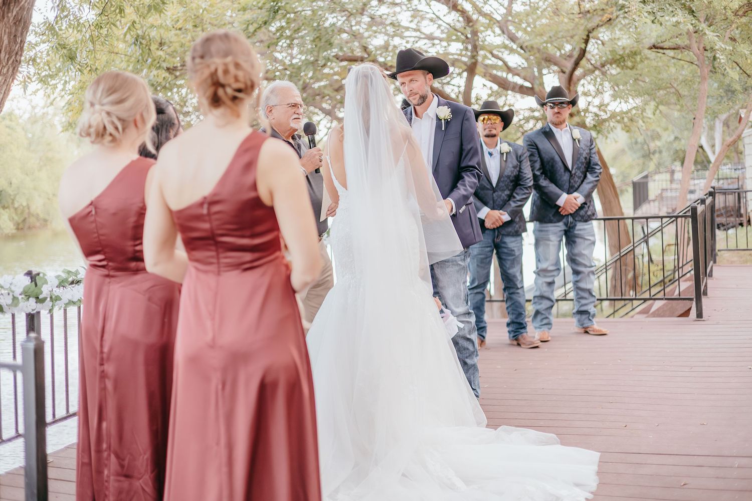 Bride in white gown and veil walks toward groom in cowboy hat at outdoor riverside wedding.