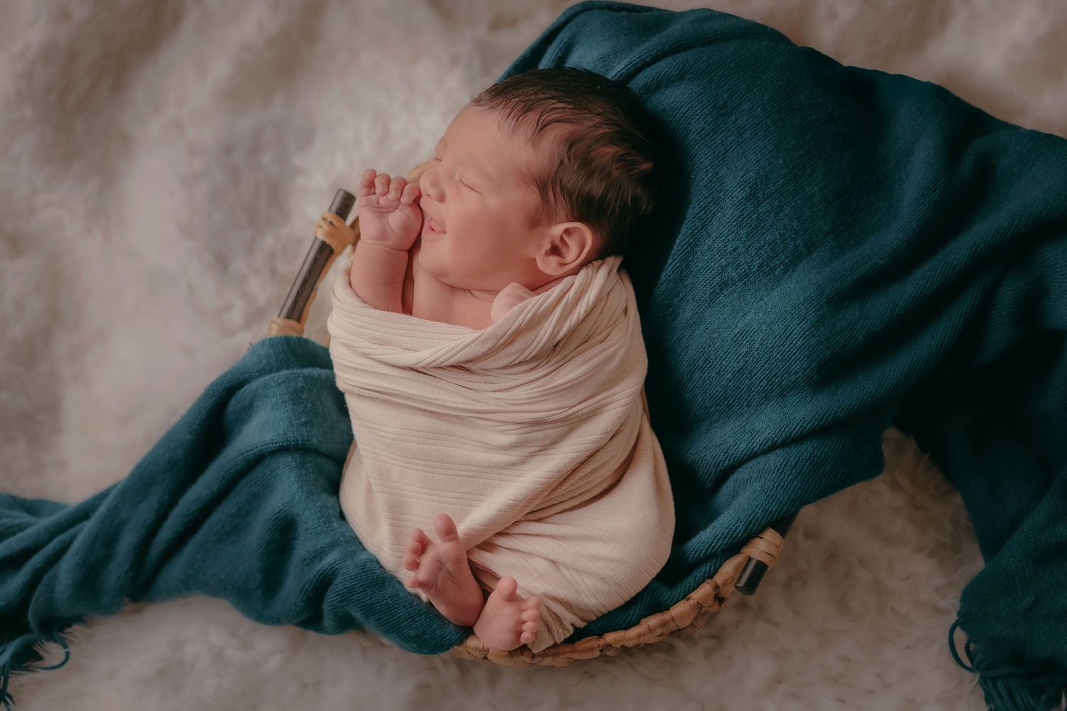 Sleeping newborn baby swaddled in cream wrap resting in basket on teal blanket.