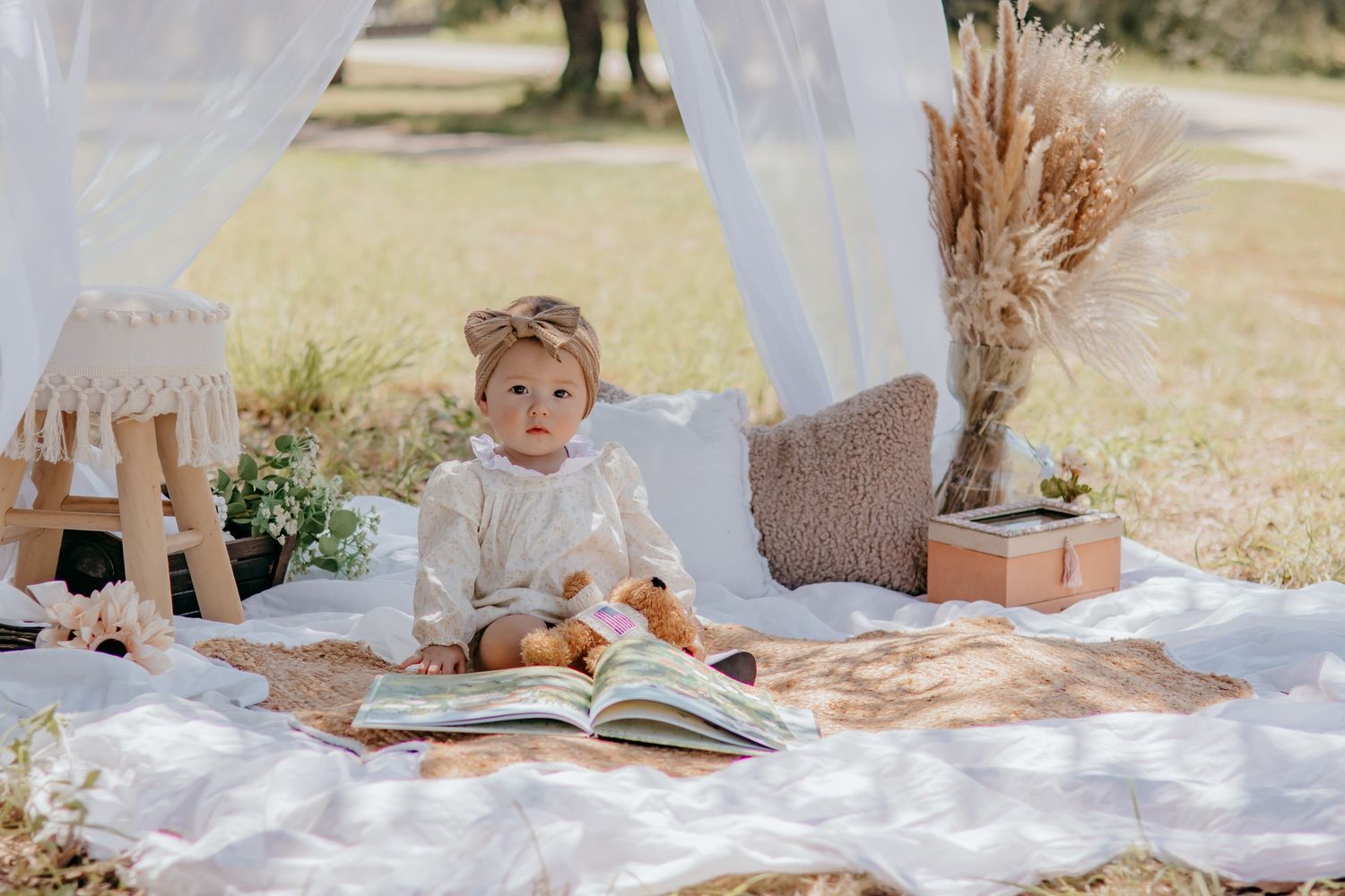 Baby girl in boho outfit sits on picnic blanket outdoors with open book, dried flowers, and canopy backdrop.