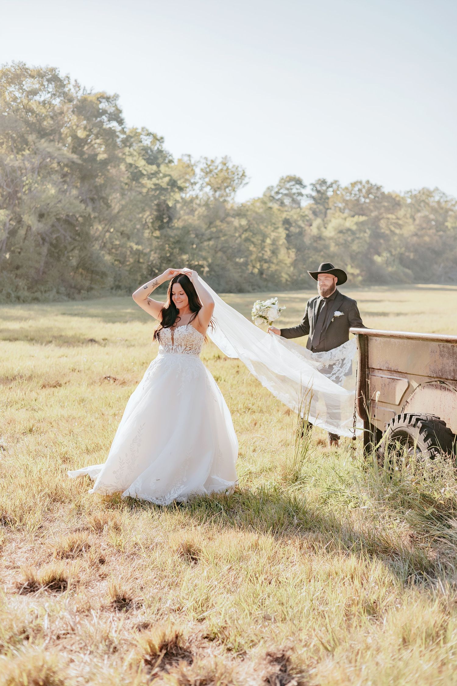 Bride in flowing white ball gown with veil poses in sunlit field near vintage rusty truck with groom.
