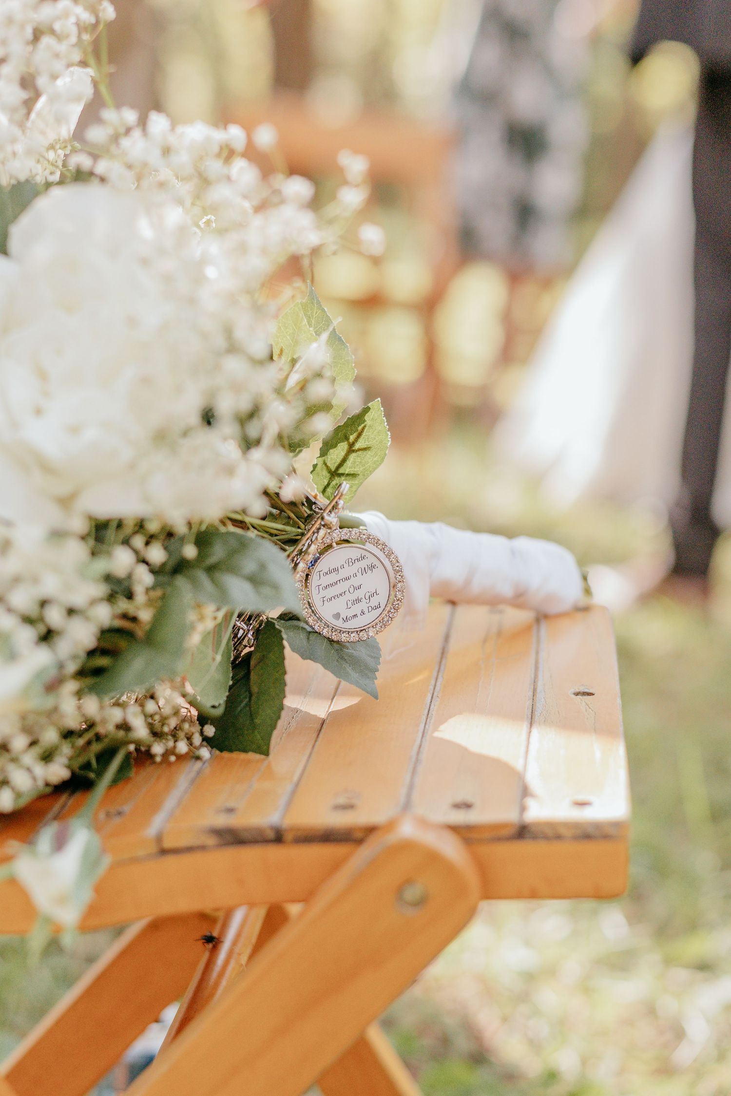 A white floral wedding bouquet with baby's breath rests on a wooden bench at an outdoor ceremony.