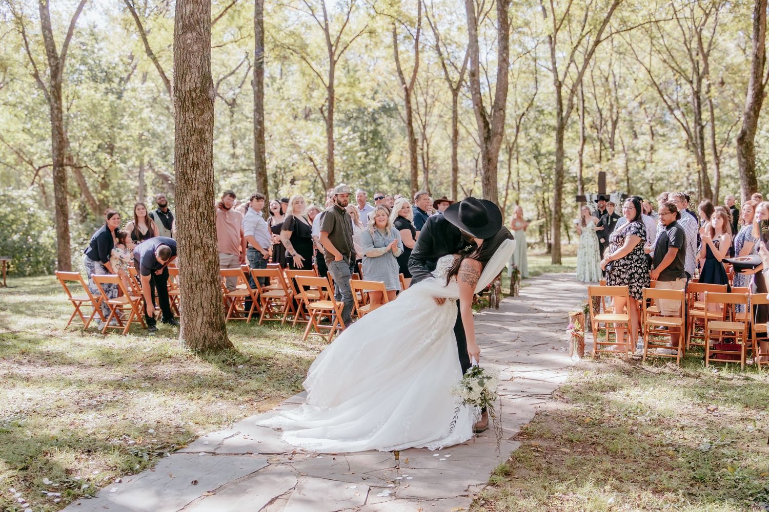 Bride and groom share a romantic dip kiss at outdoor woodland wedding ceremony with guests seated on wooden chairs.