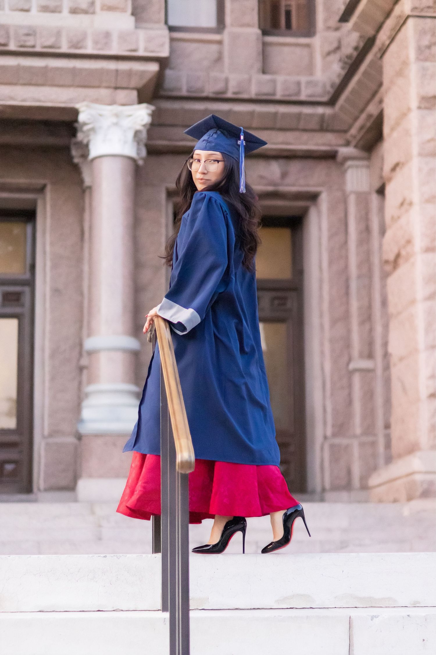 Graduate in navy cap and gown poses on steps of stone building in red dress.