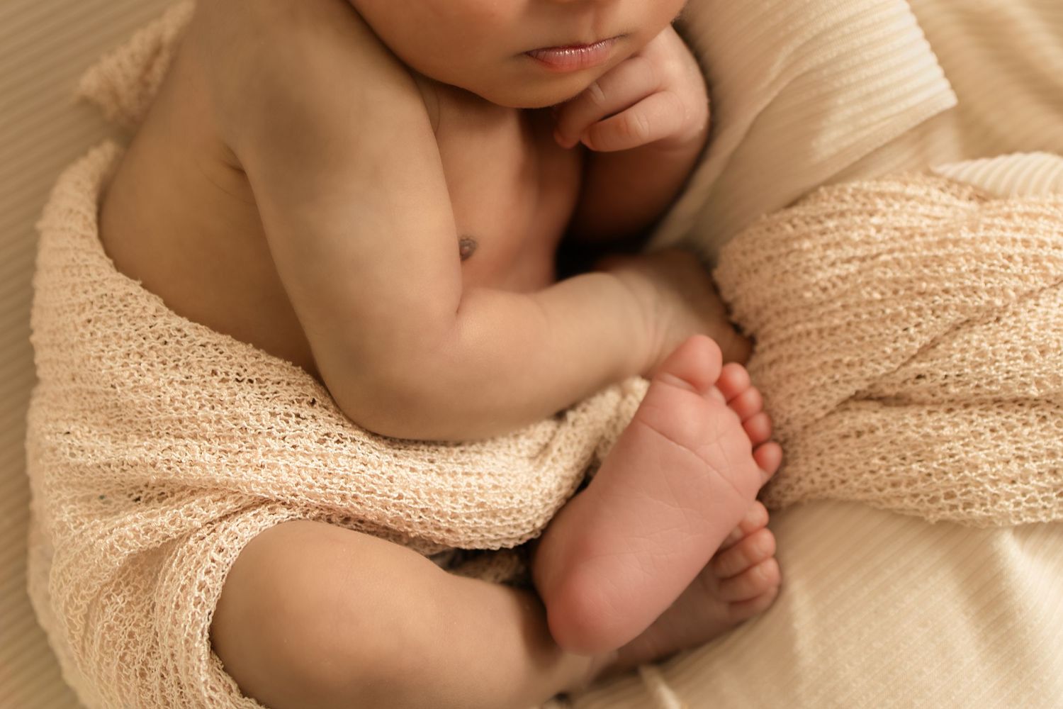 Close-up of a peaceful newborn baby wrapped in a soft beige knit blanket, showing tiny feet and hands.