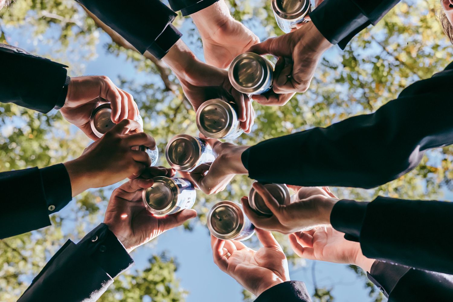 Group of people in formal attire toasting with glasses outdoors, viewed from below with trees in background.