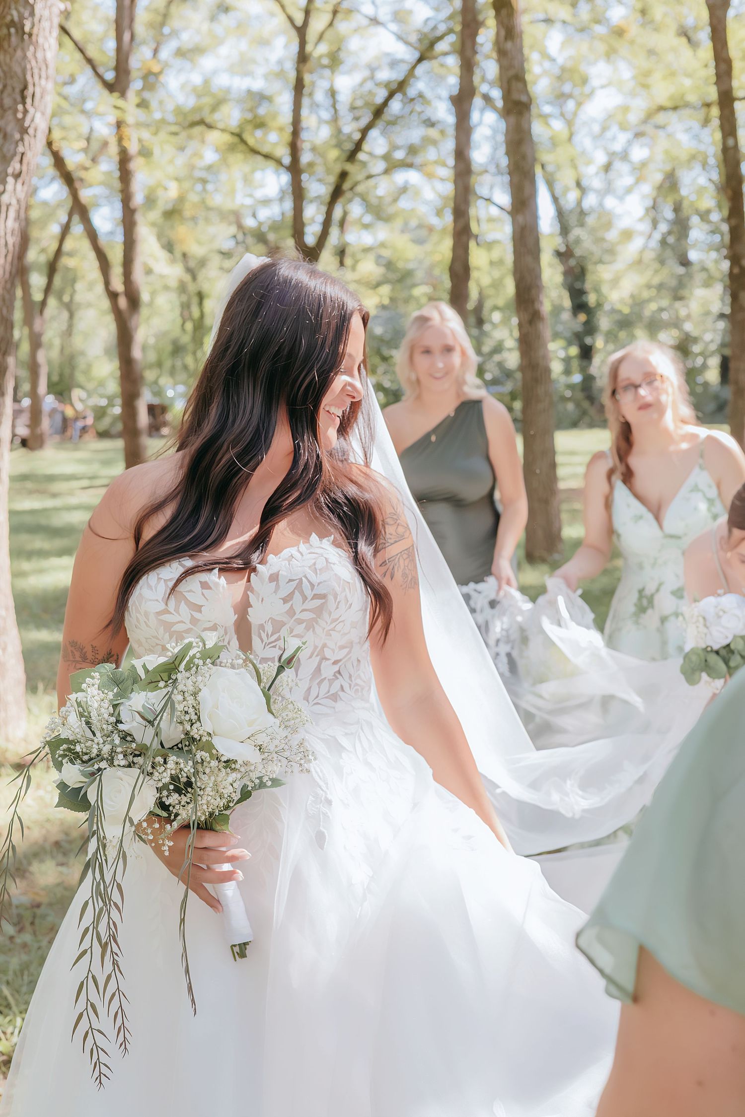Bride in white lace gown holds greenery bouquet outdoors, bridesmaids in sage green dresses carry veil behind her.