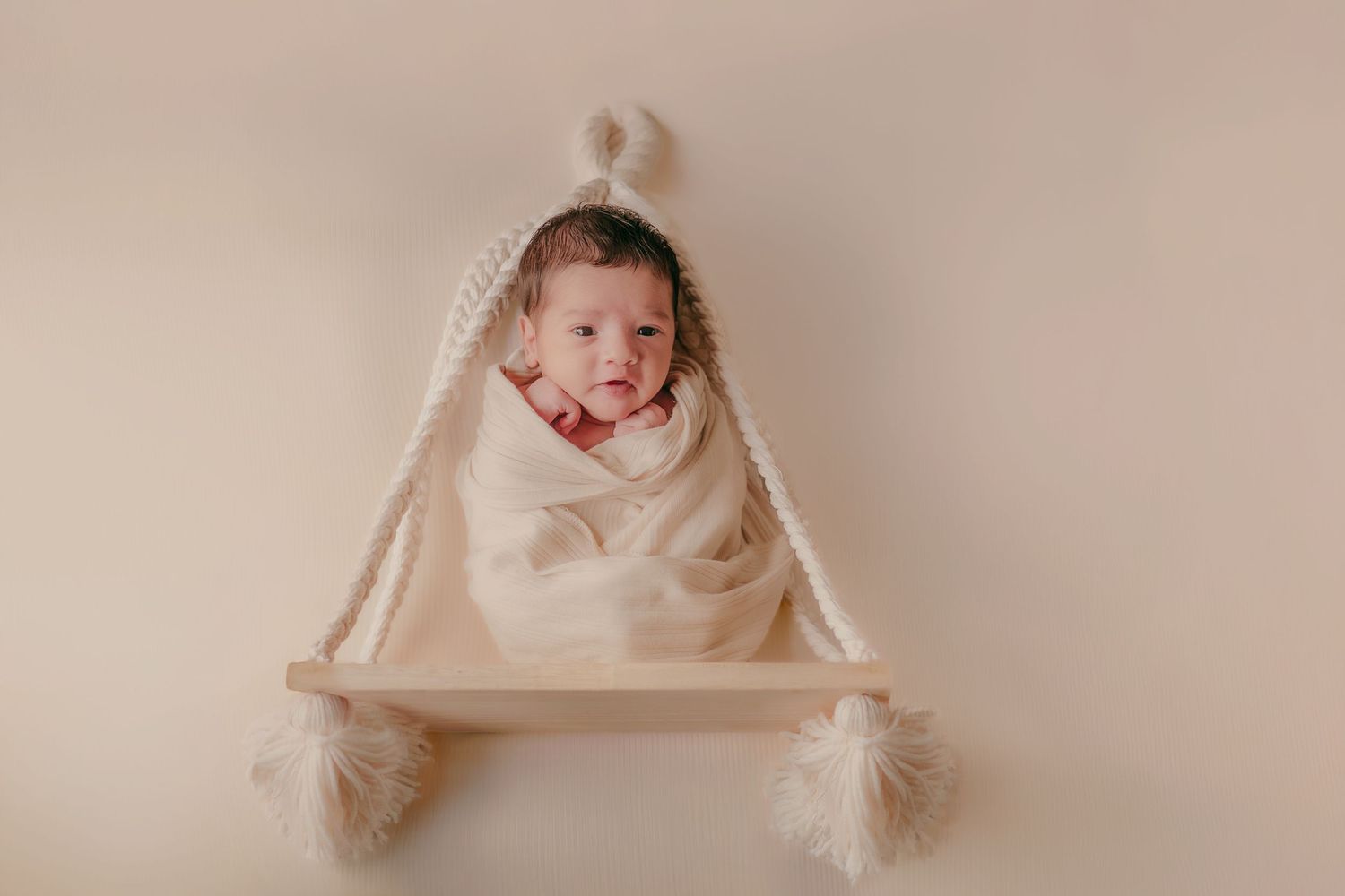 Awake newborn baby wrapped in cream swaddle wearing knit bunny ear hat on wooden shelf.