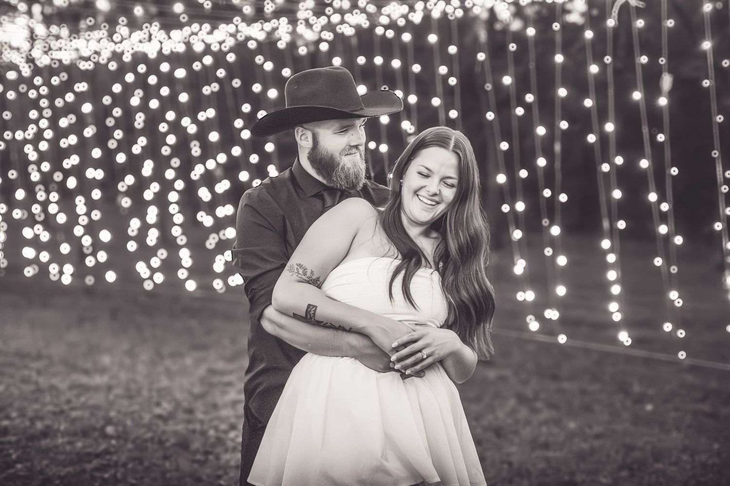 Black and white photo of a couple embracing in front of a sparkling fairy light backdrop outdoors.