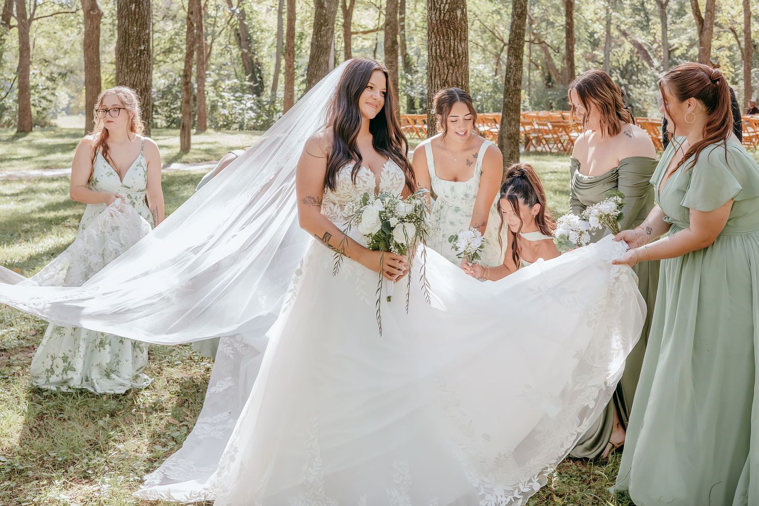 Bridesmaids in sage green dresses help adjust bride's white gown and veil in sunlit forest setting.
