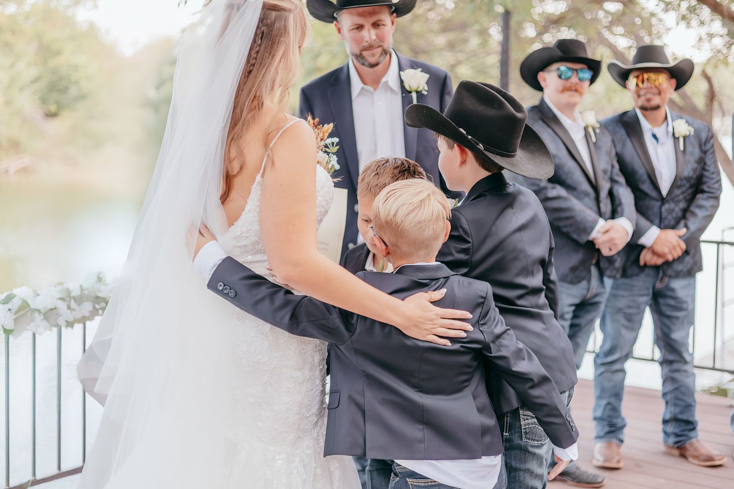 Bride being hugged by children in cowboy hats during outdoor wedding ceremony.