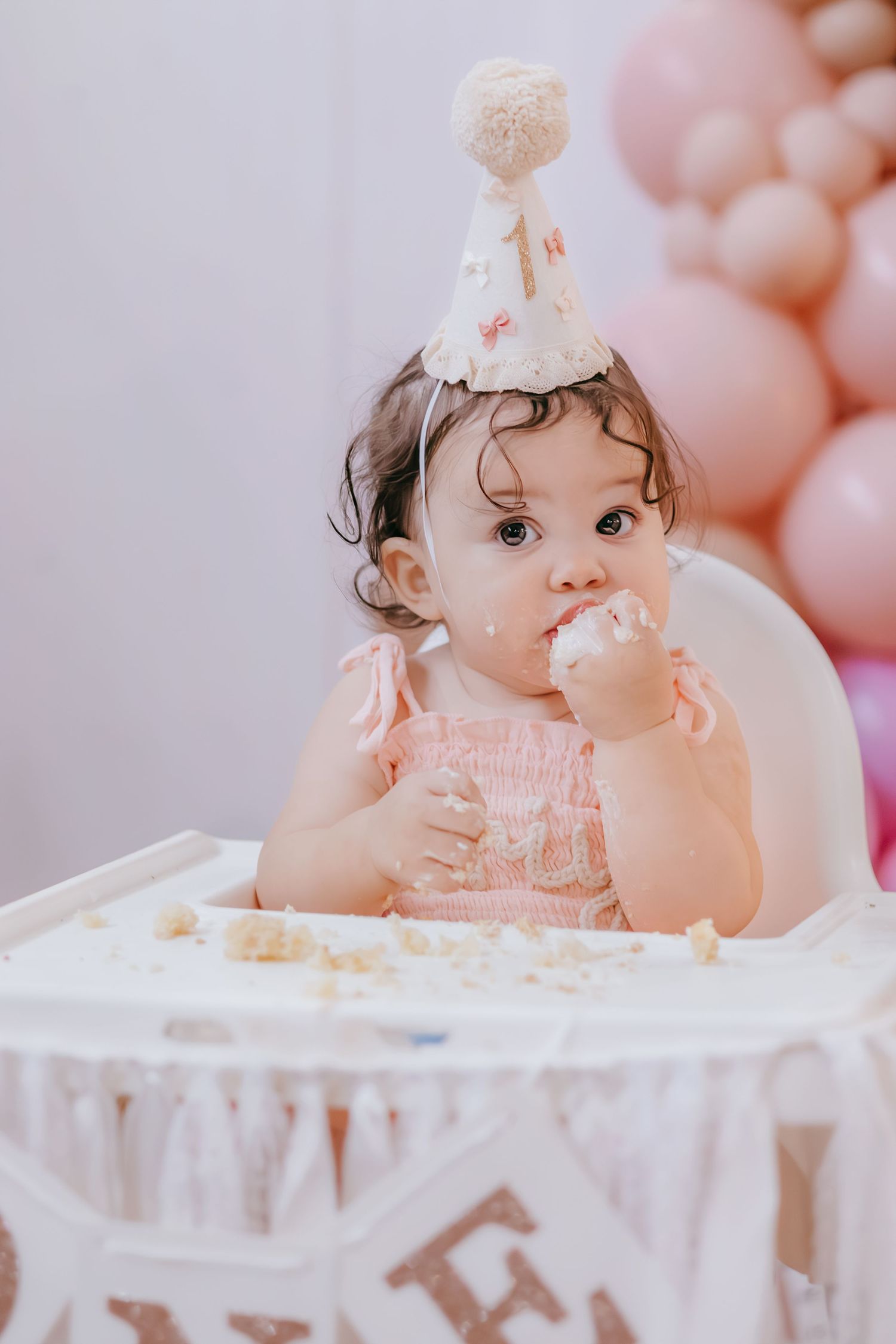 Baby girl in pink dress and party hat smashes first birthday cake in high chair with pink balloon backdrop.