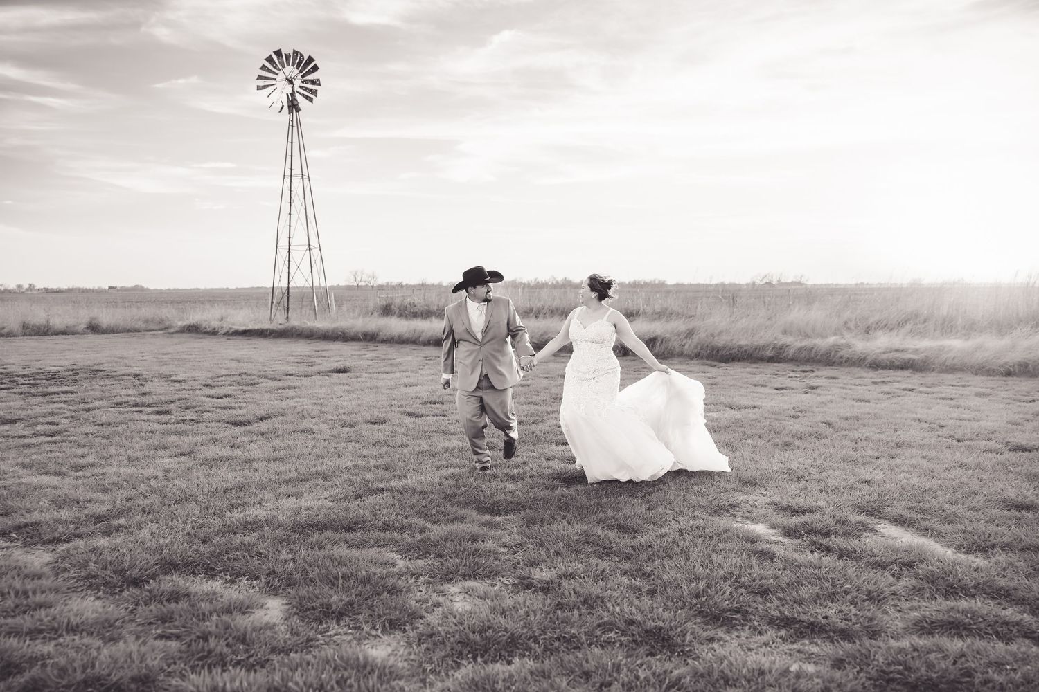 Black and white photo of bride and groom running hand in hand through a field with a windmill backdrop.