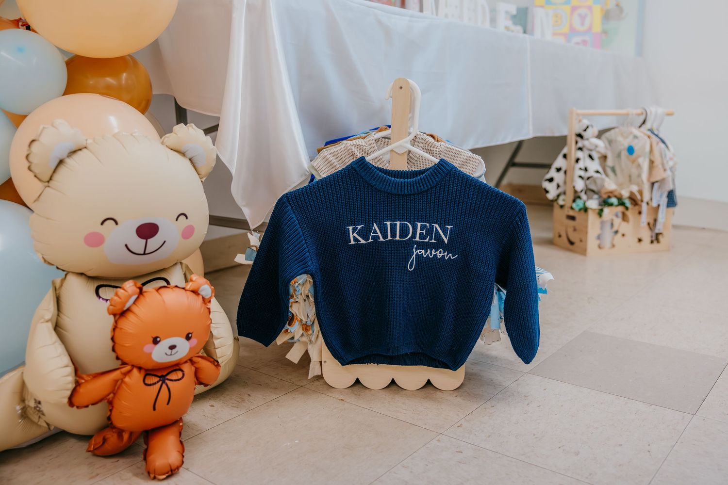 Navy blue personalized baby sweatshirt reading KAIDEN hung on wooden hanger beside teddy bear balloons at a baby shower.
