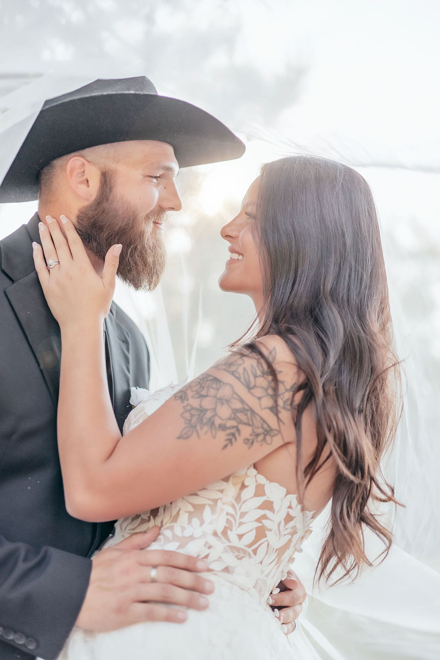 A smiling bearded groom in a black cowboy hat embraces his tattooed bride in a lace wedding dress under her veil.
