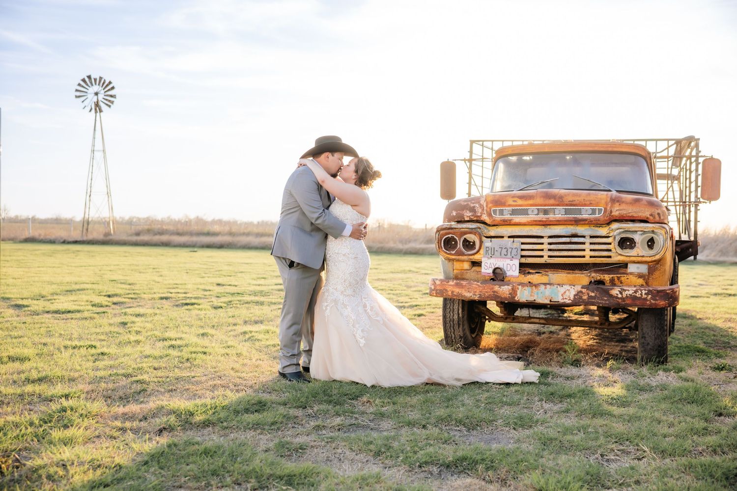 Bride and groom kissing beside a rustic orange vintage truck in a farm field with a windmill backdrop.