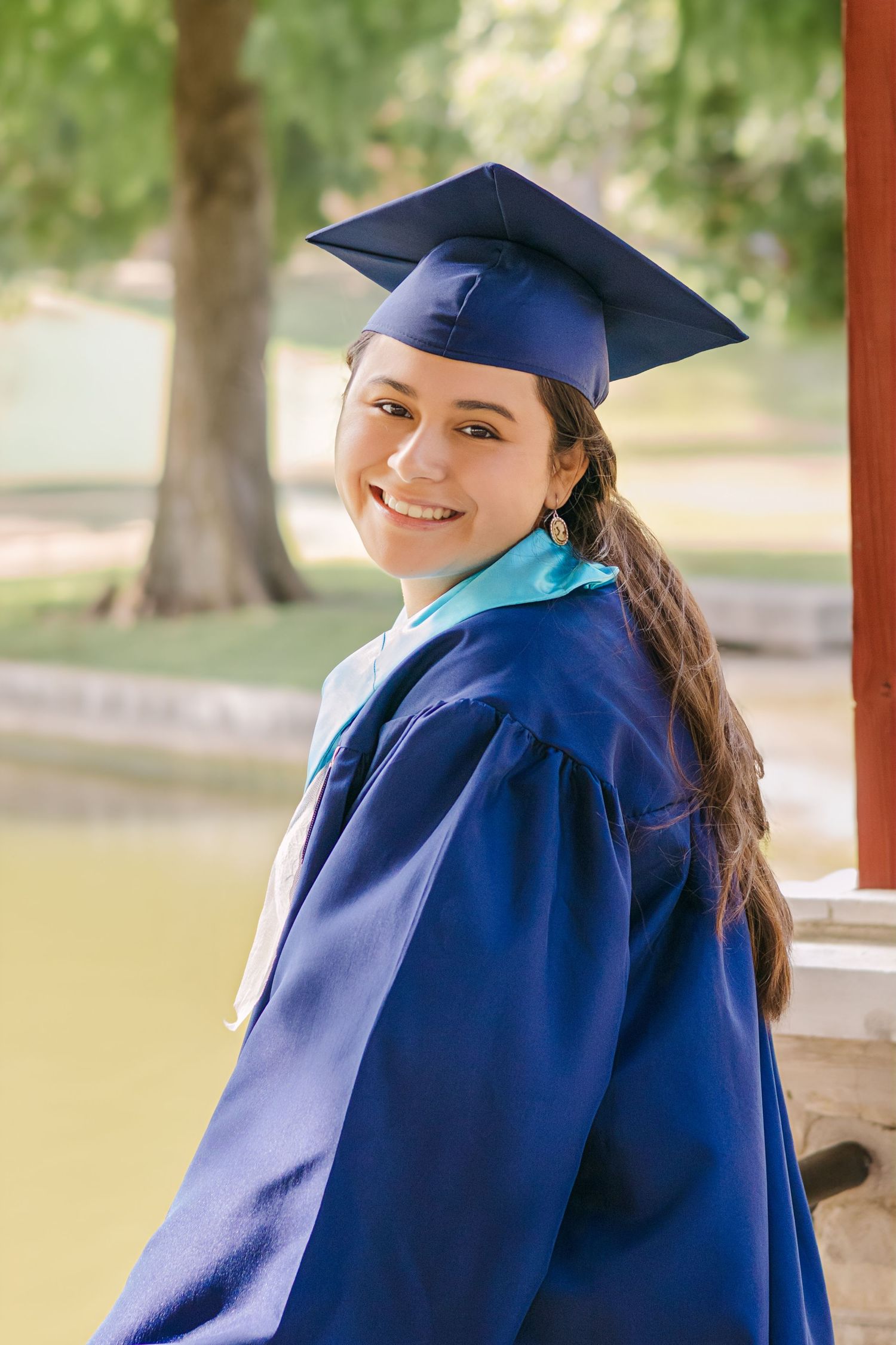 Smiling young woman in blue graduation cap and gown posing outdoors near a pond in a park.