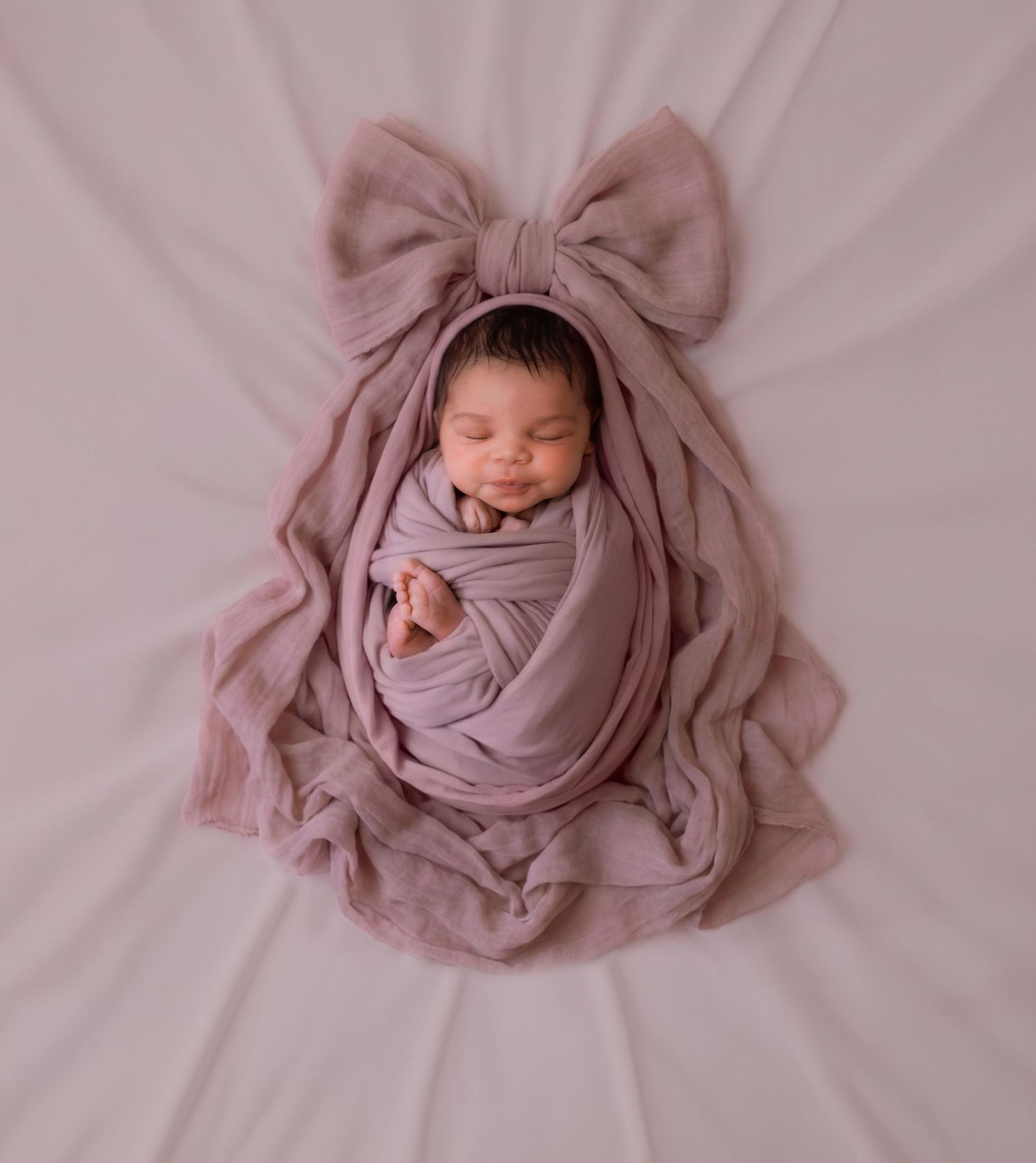 Sleeping newborn baby wrapped in purple swaddle with matching bow headband on pink background.