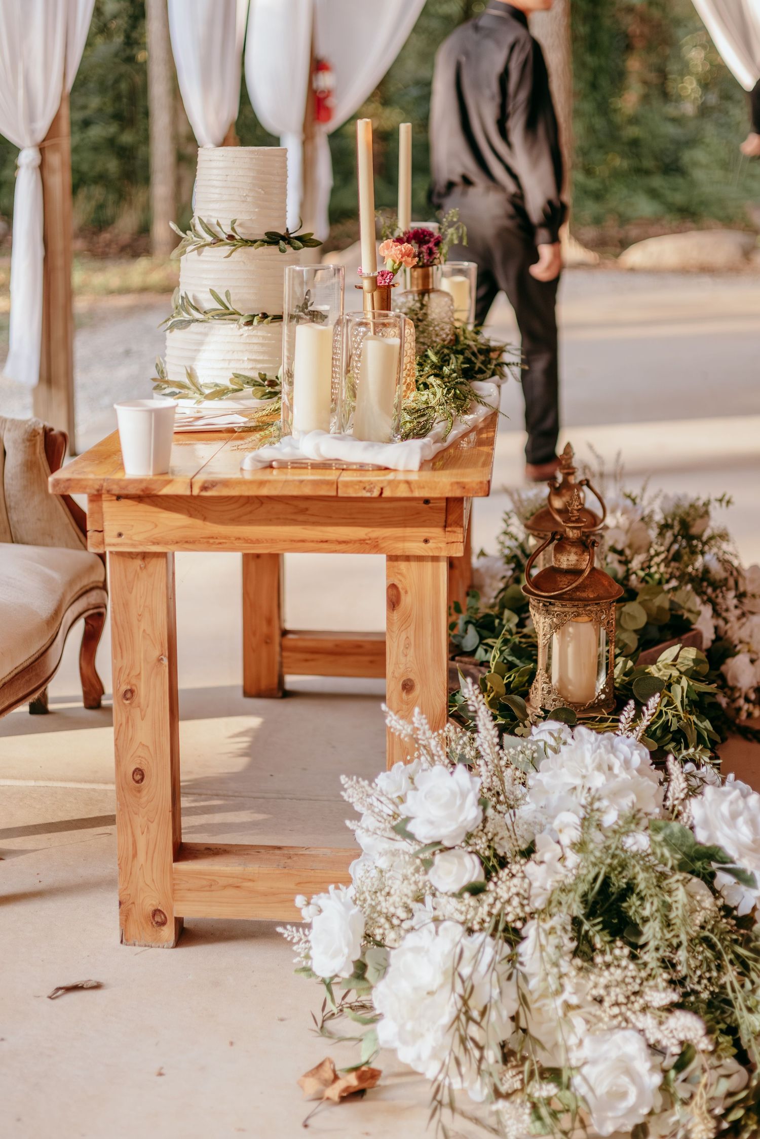 Rustic wooden table with white wedding cake, candles, and greenery surrounded by white floral arrangements at an outdoor wedding.
