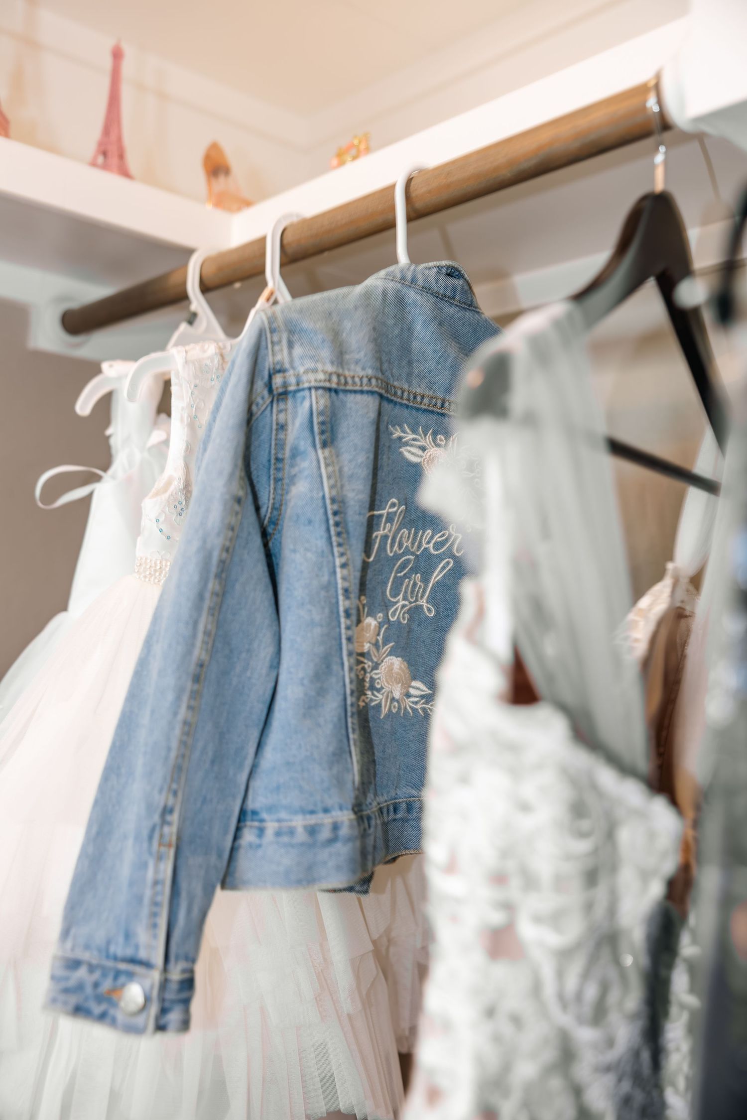 Custom embroidered flower girl denim jacket hanging on a rack beside white tulle wedding dresses.