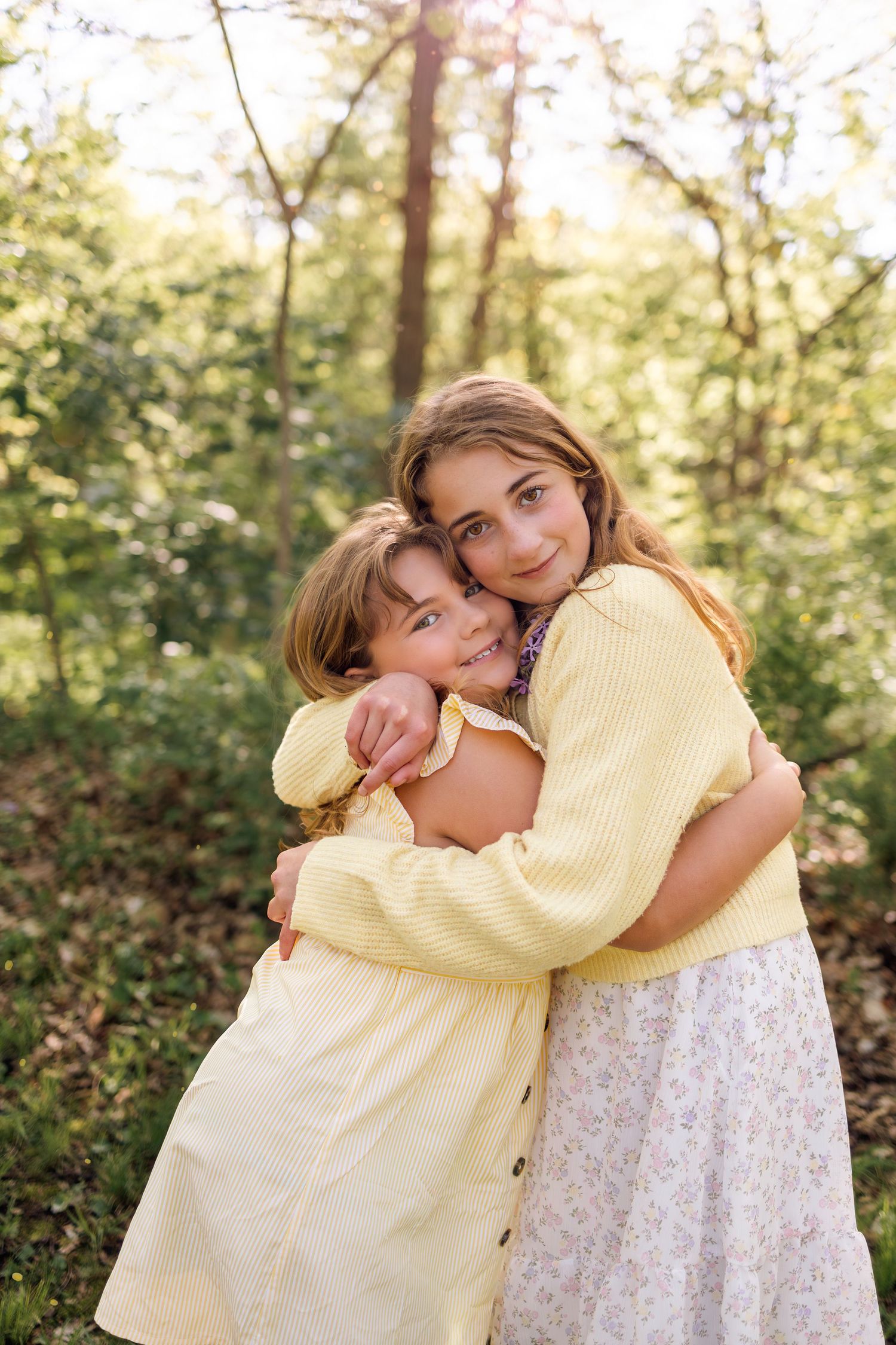 Two young girls hugging warmly in a sunlit forest, wearing yellow outfits, smiling joyfully outdoors.