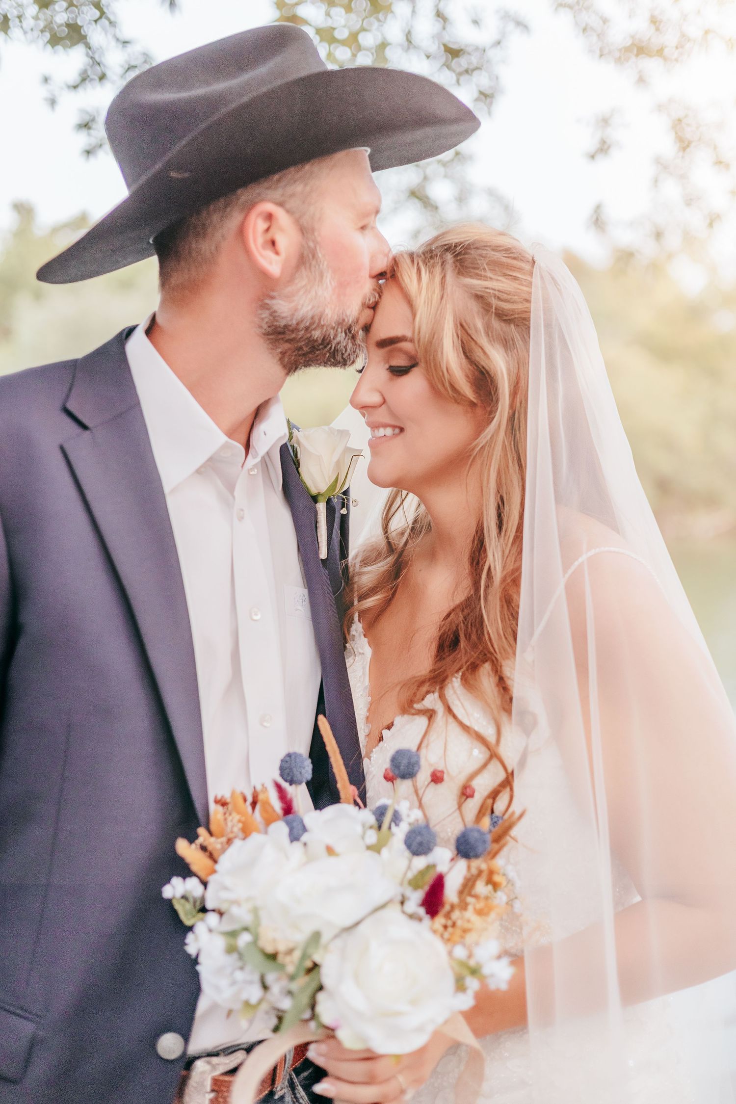 Groom in cowboy hat kisses bride on forehead as she smiles holding white floral bouquet outdoors.