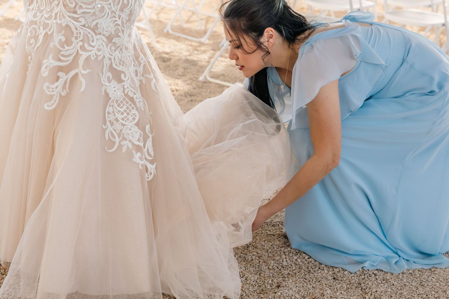 Bridesmaid in blue dress adjusting the hem of a white lace wedding gown outdoors. Taylor, Texas