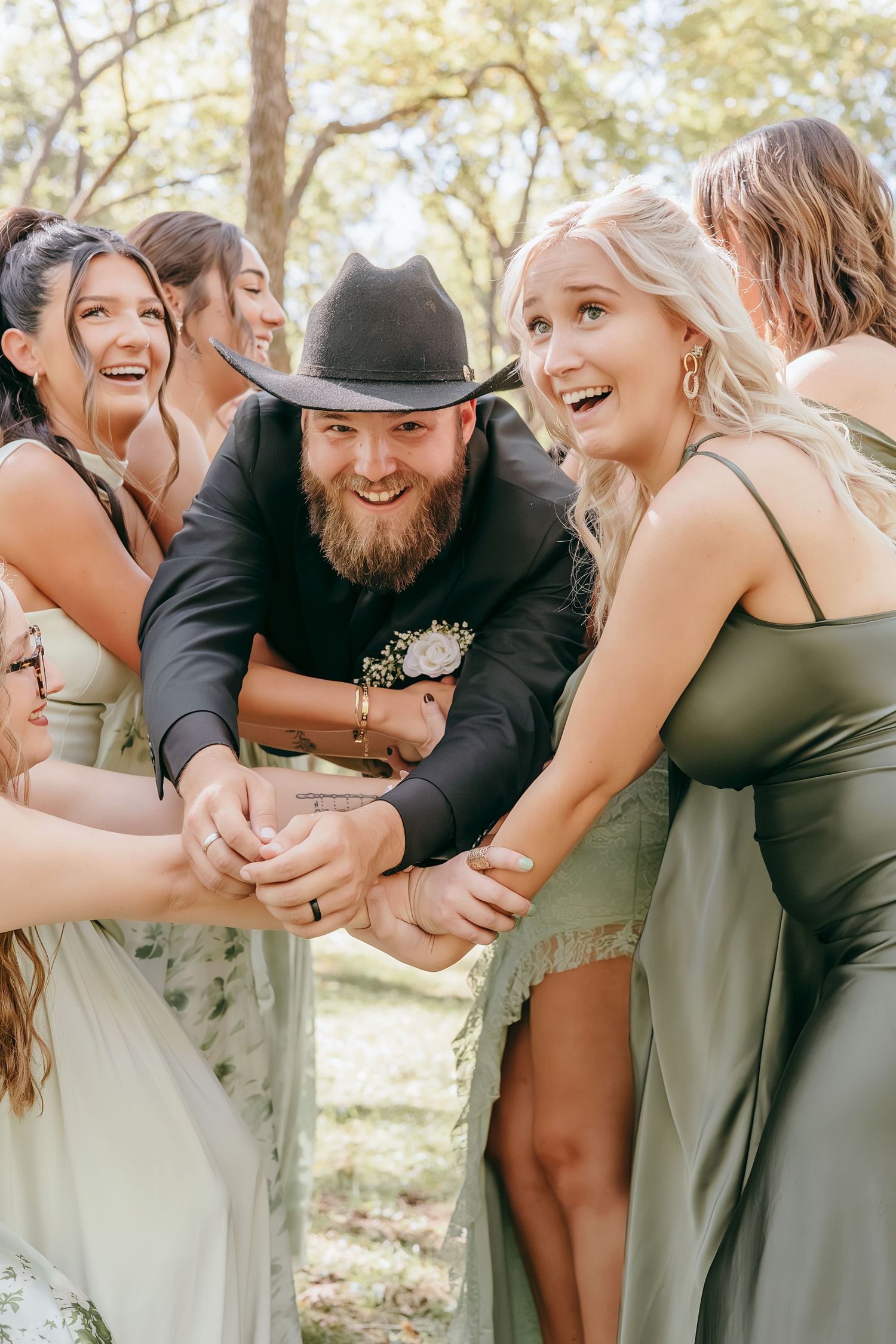 Bearded groom in black cowboy hat laughing and holding hands with bridesmaids in sage green dresses outdoors.