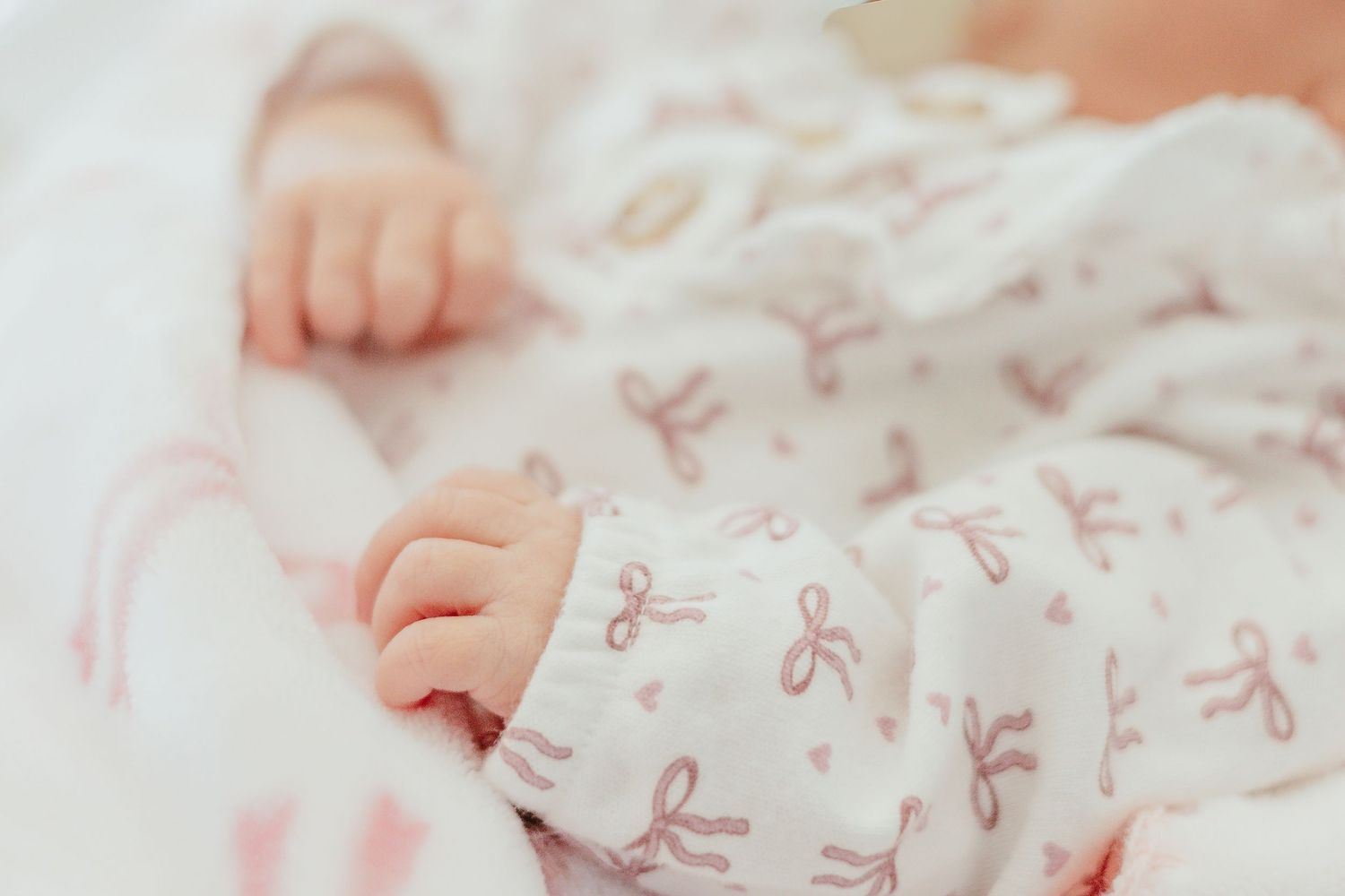Close-up of a newborn baby in a white onesie with pink bow print, showing tiny delicate hands and fingers.