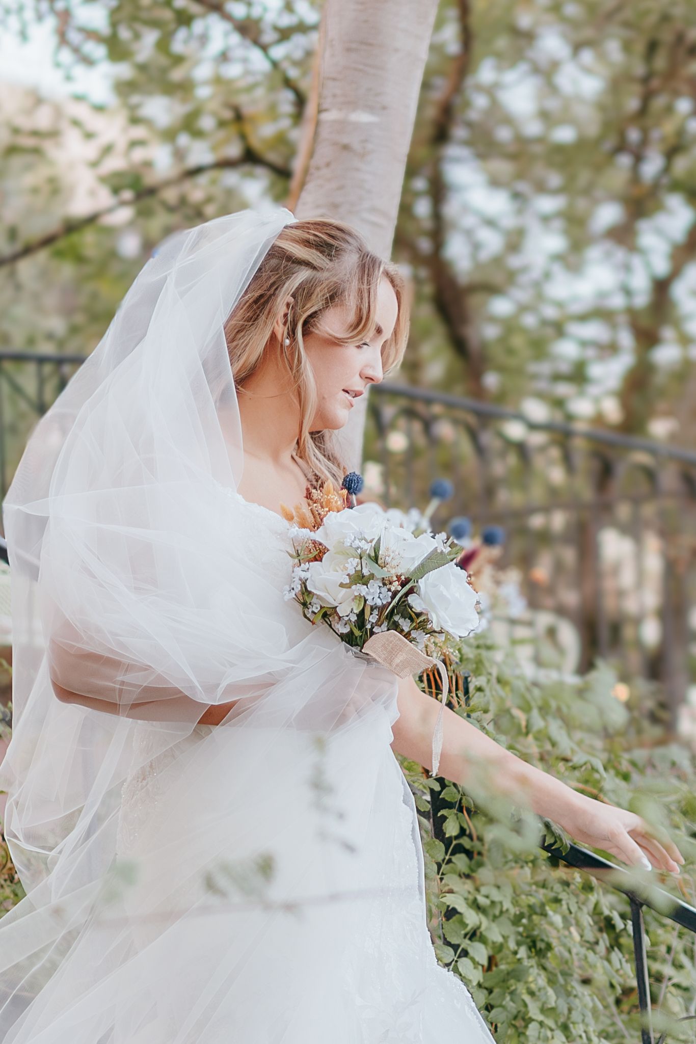 Bride in white veil and gown holding white floral bouquet on outdoor staircase.