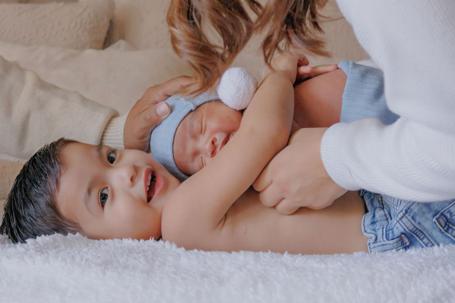 Older toddler in blue hat lies beside sleeping newborn sibling while mother gently holds them on white blanket.