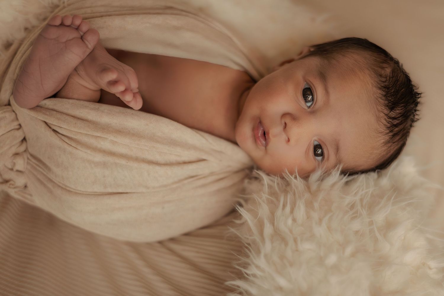 Awake newborn baby wrapped in cream swaddle cloth resting on a fluffy white fur blanket.