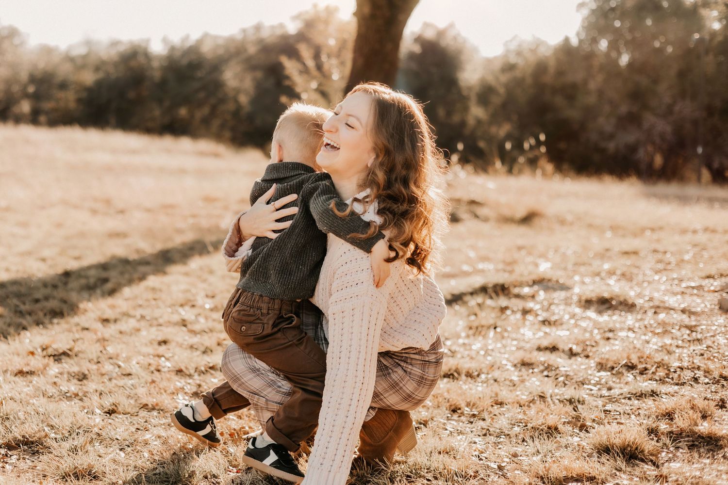 A young mother kneeling in a sunlit field hugging and laughing with her toddler son.