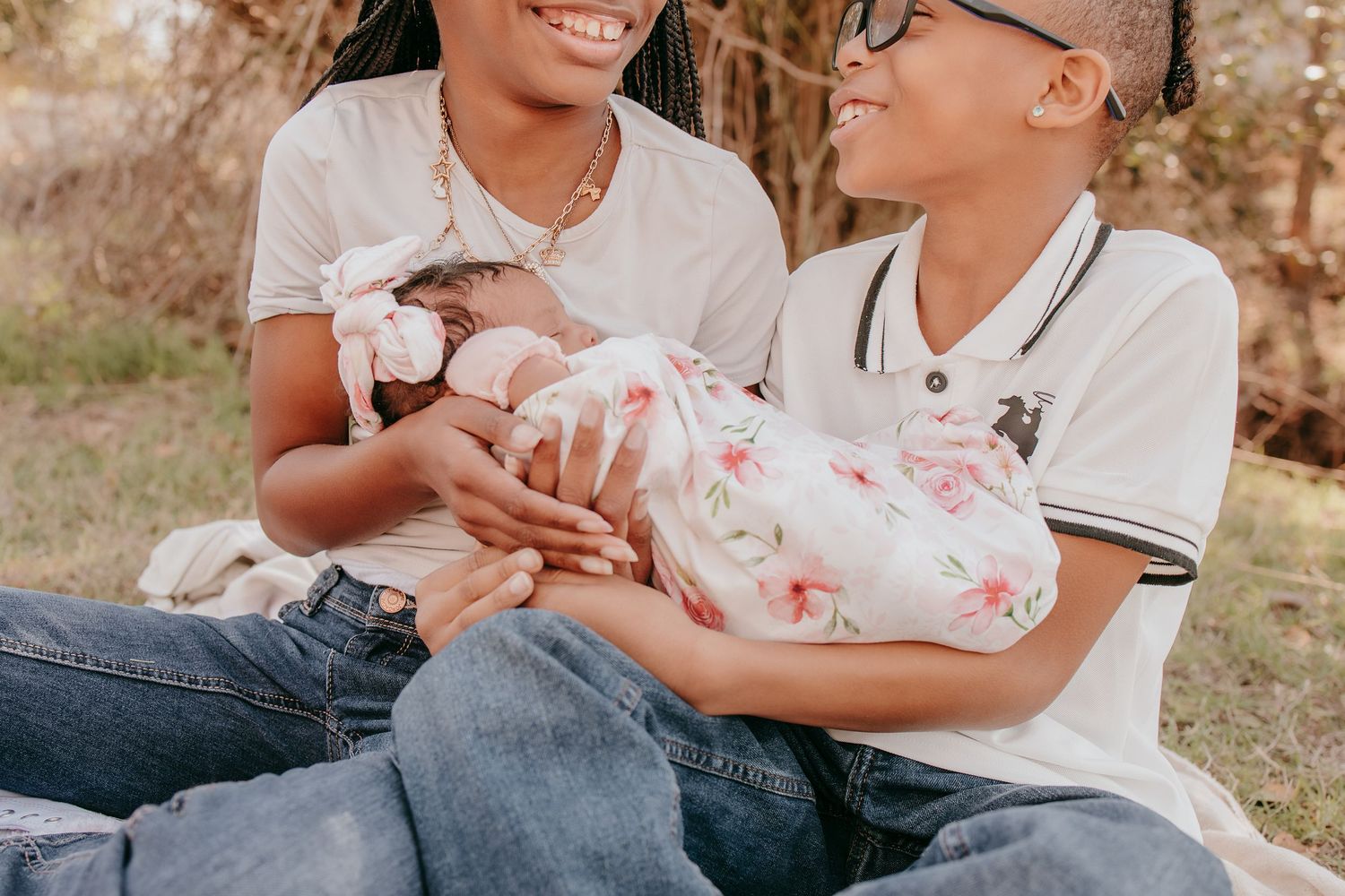Smiling mother with braids holding newborn baby in floral swaddle with older child beside her outdoors.