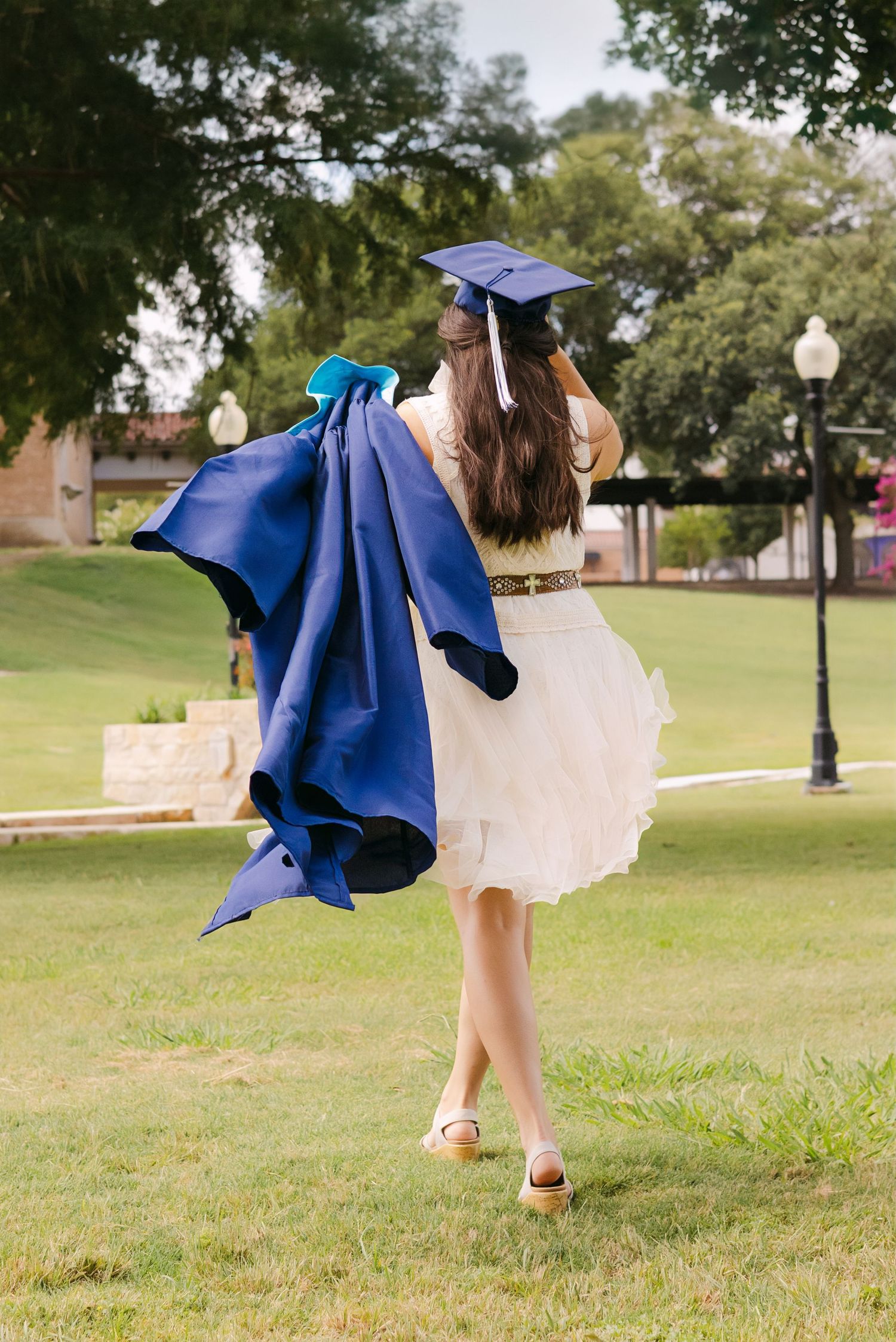 Graduate in white dress carries blue cap and gown across sunny green campus lawn.