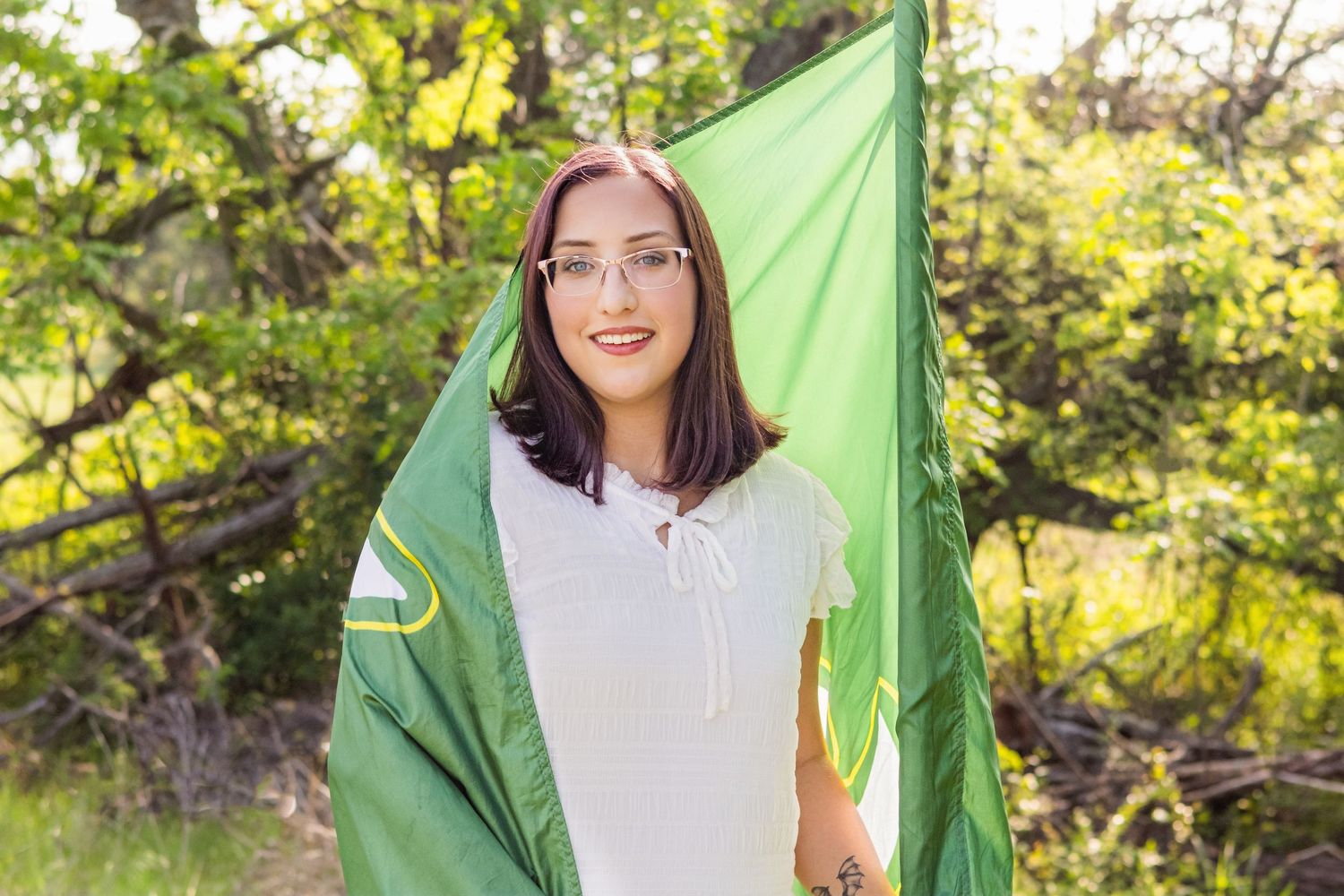 Senior Color Guard girl with glasses smiling outdoors, holding a green flag surrounded by lush green trees.