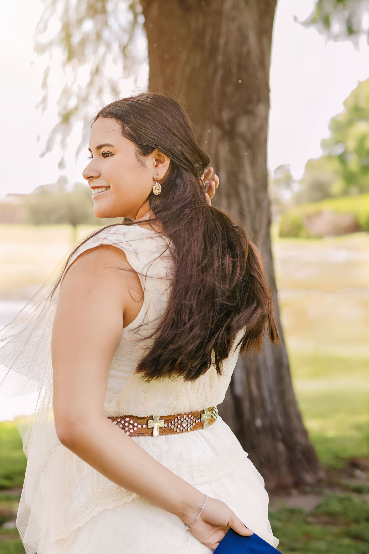 Smiling brunette woman in white dress with gold belt stands by large tree in sunny outdoor setting.