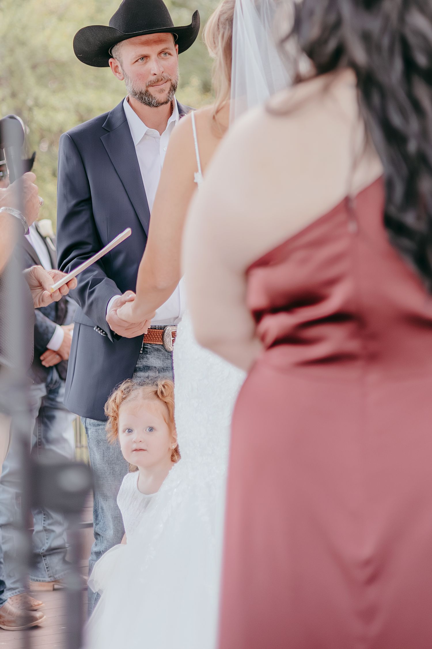 Groom in black cowboy hat reads vows as flower girl watches during outdoor wedding ceremony.