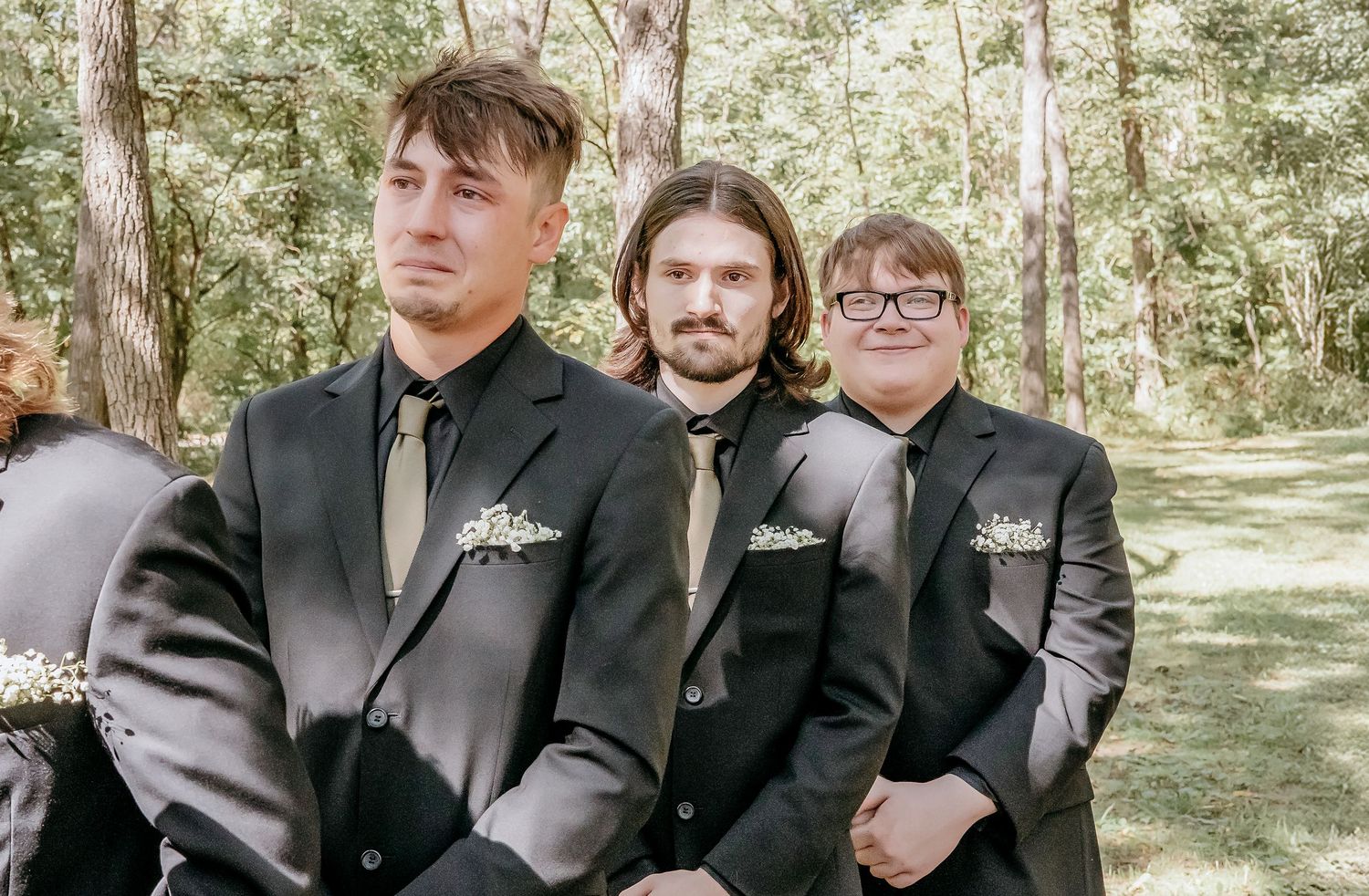 Three groomsmen in dark suits with floral boutonnieres seated outdoors at a wedding ceremony.