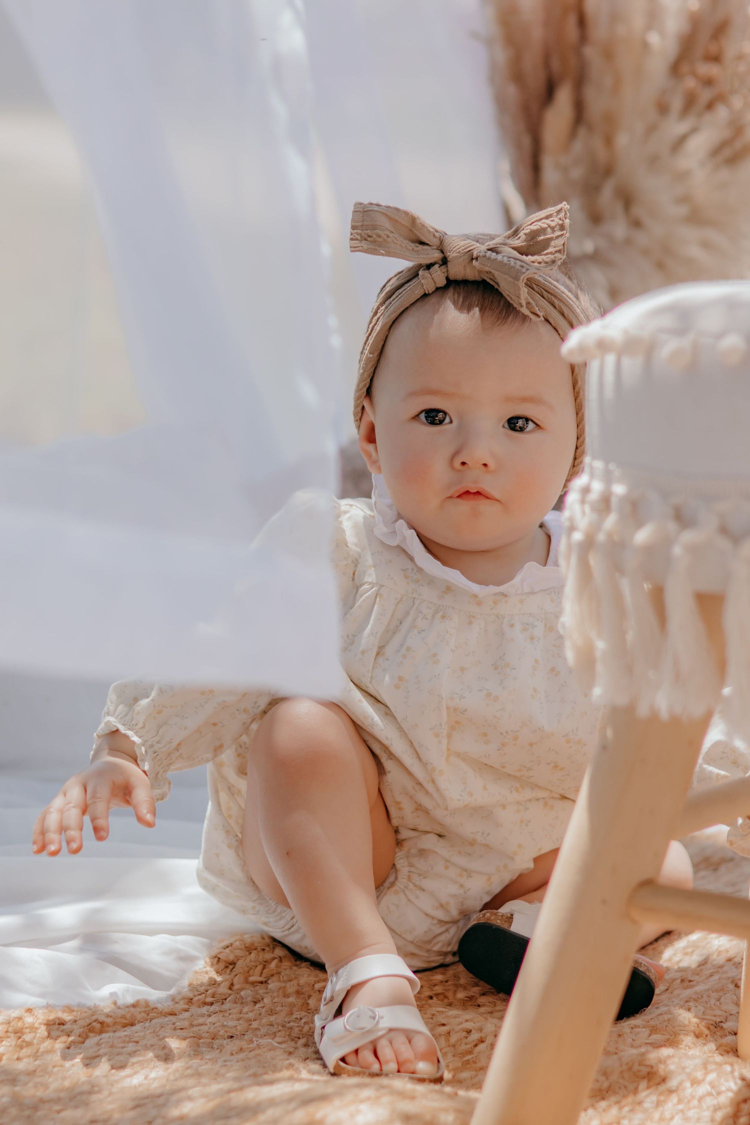 Adorable baby girl sitting on a woven rug wearing a floral romper and beige bow headband in a boho setting.