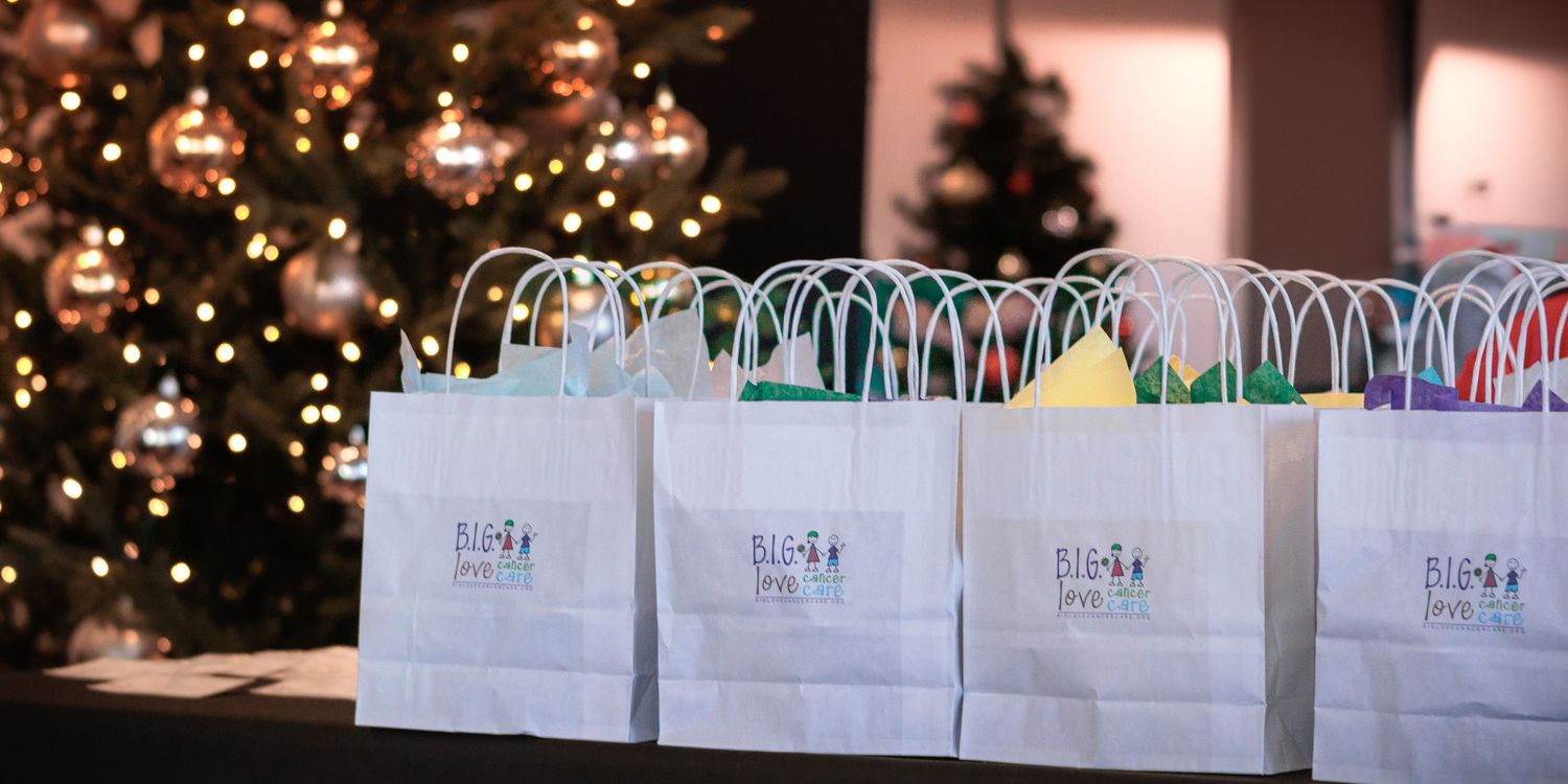 Rows of white gift bags with colorful tissue paper arranged on a table in front of a decorated Christmas tree.