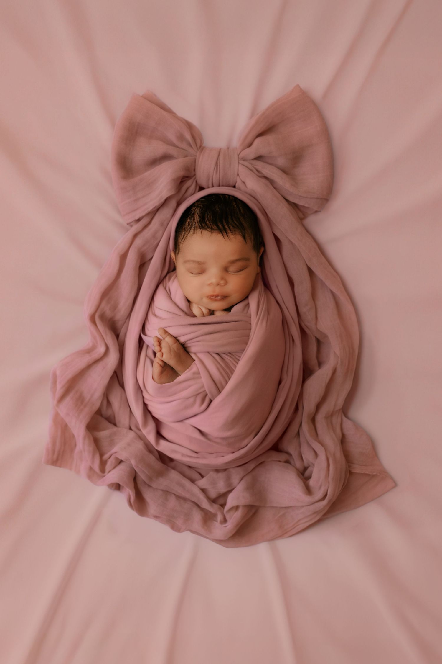 A peacefully sleeping newborn is wrapped in a soft pink swaddle with a large bow on a pink background.
