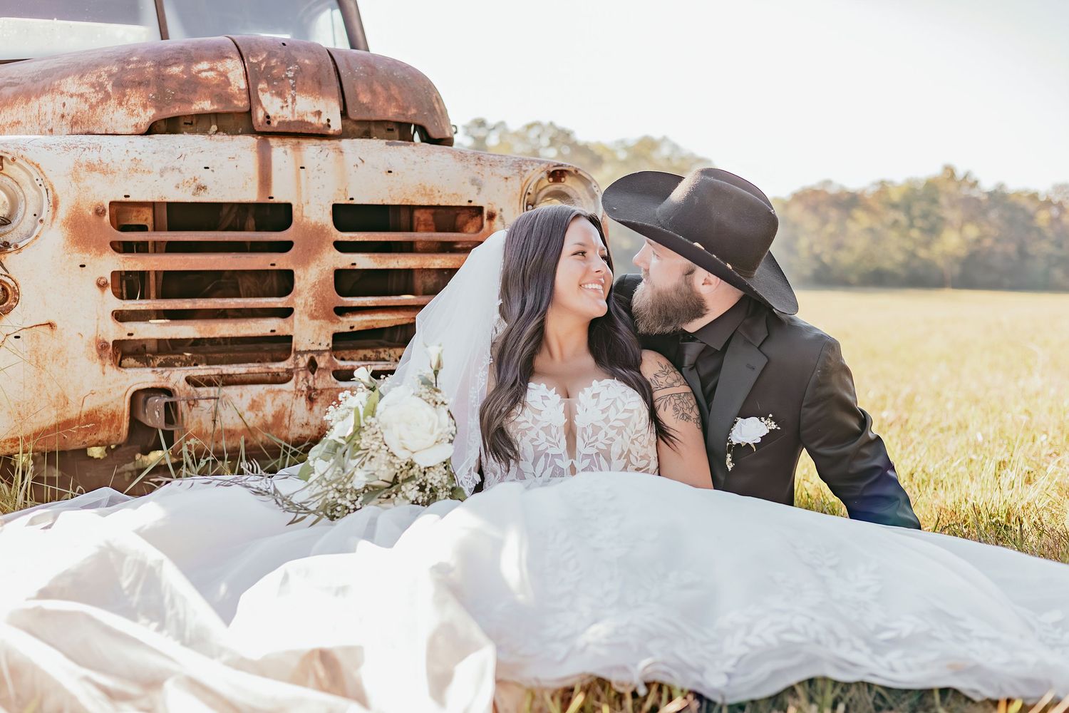 Bride and bearded groom in cowboy hat sitting together in field beside a rustic vintage red truck.