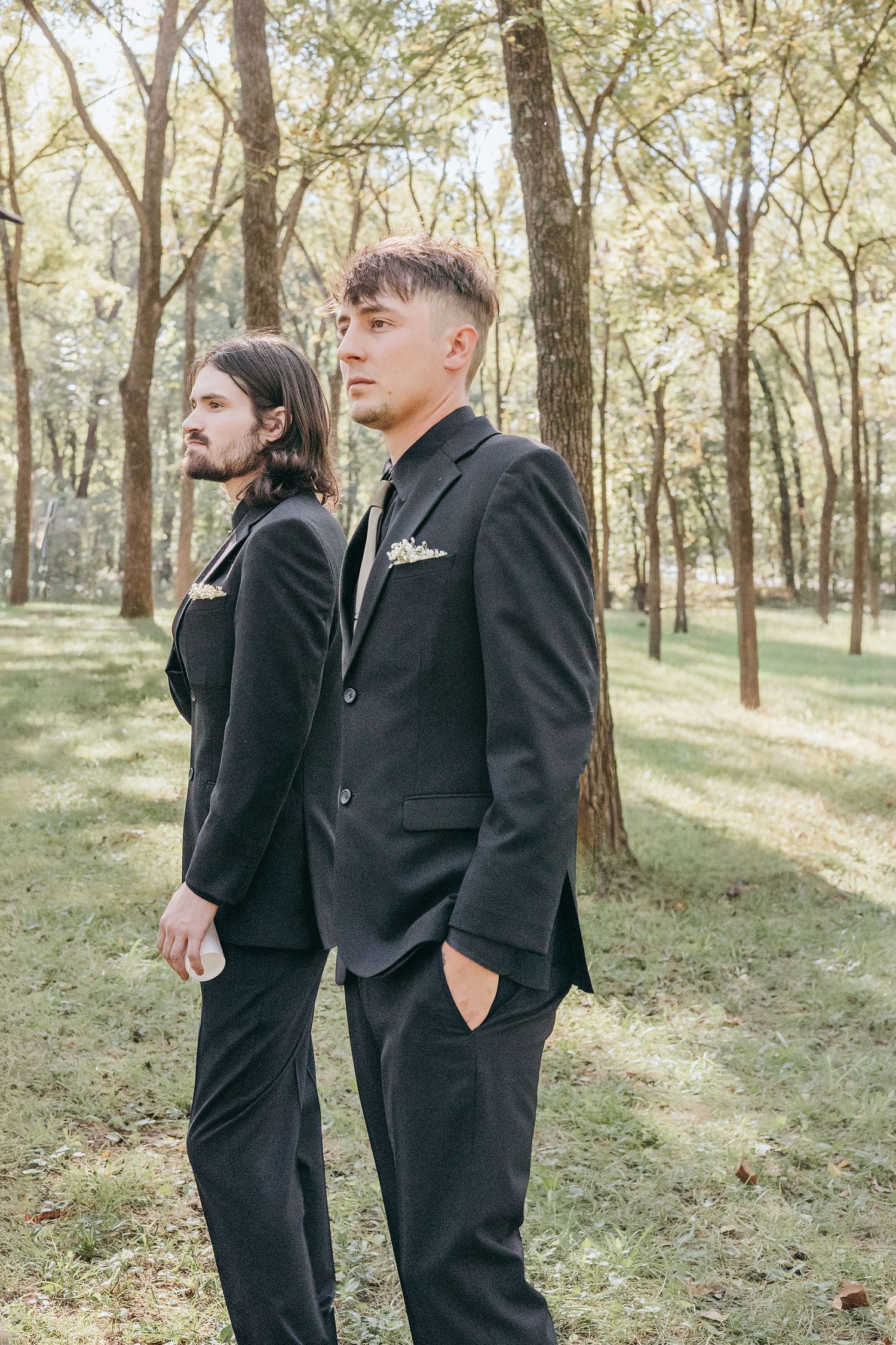 Two groomsmen in matching black suits stand back-to-back in a sunlit wooded outdoor setting.